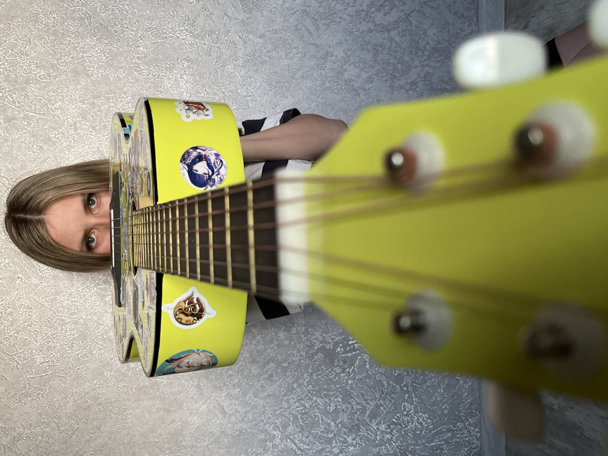 A woman is holding a yellow acoustic guitar, looking at the camera. The guitar is adorned with anime stickers. She is wearing a black and white striped top. The location appears to be indoors with a textured, light-colored wall. The image is likely taken in portrait orientation with the camera close to the guitar, creating a blurred perspective. There is no verifiable information to confirm the location as Chaikovsky, Russia.