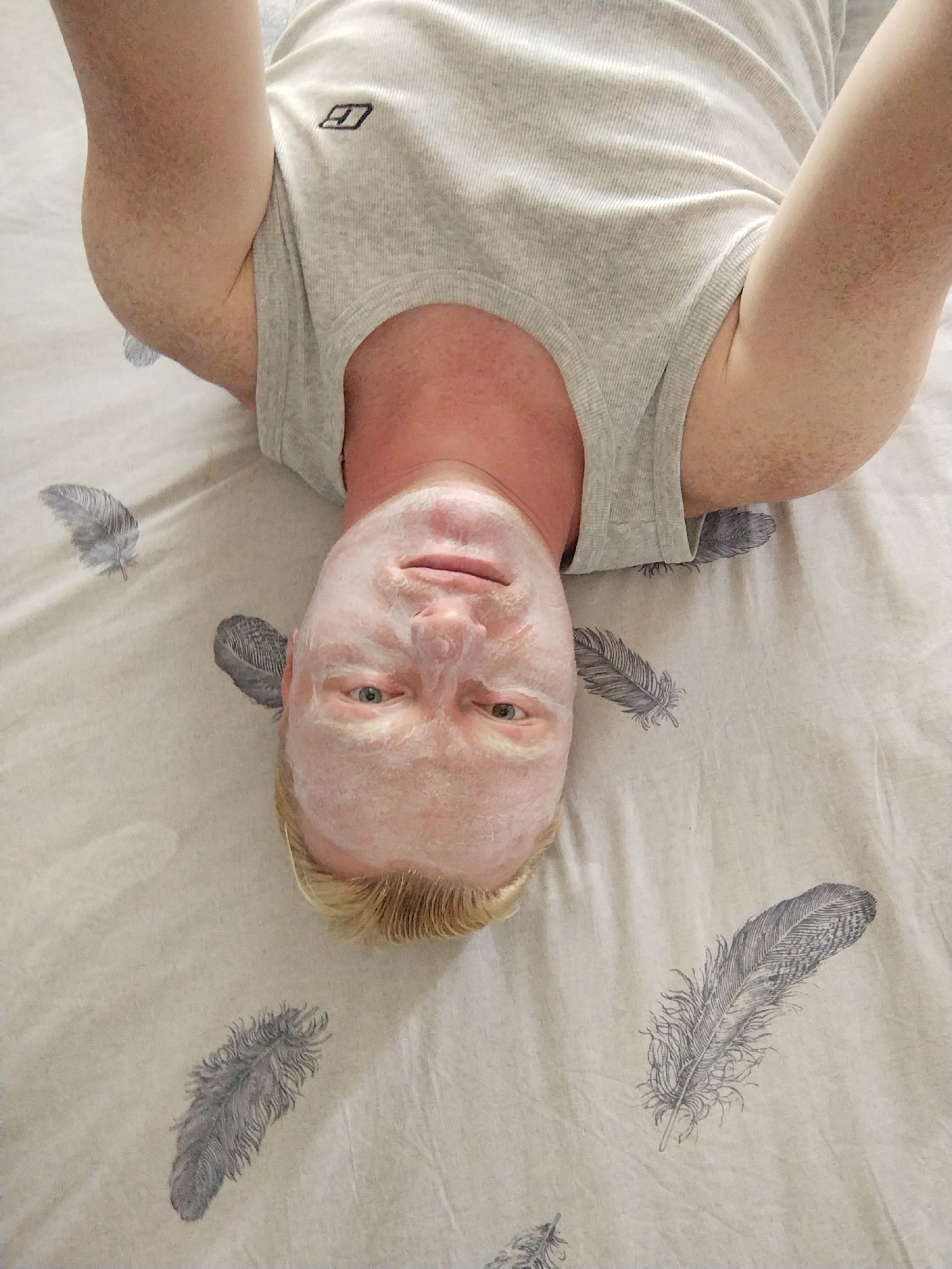 A person is lying on their back on a bed with a white sheet patterned with grey feathers. The person is wearing a light grey ribbed tank top. Their face is covered in a white facial mask. Their eyes are open and looking upwards. Their arms are raised above their head. The overall scene appears to be in a domestic indoor setting.