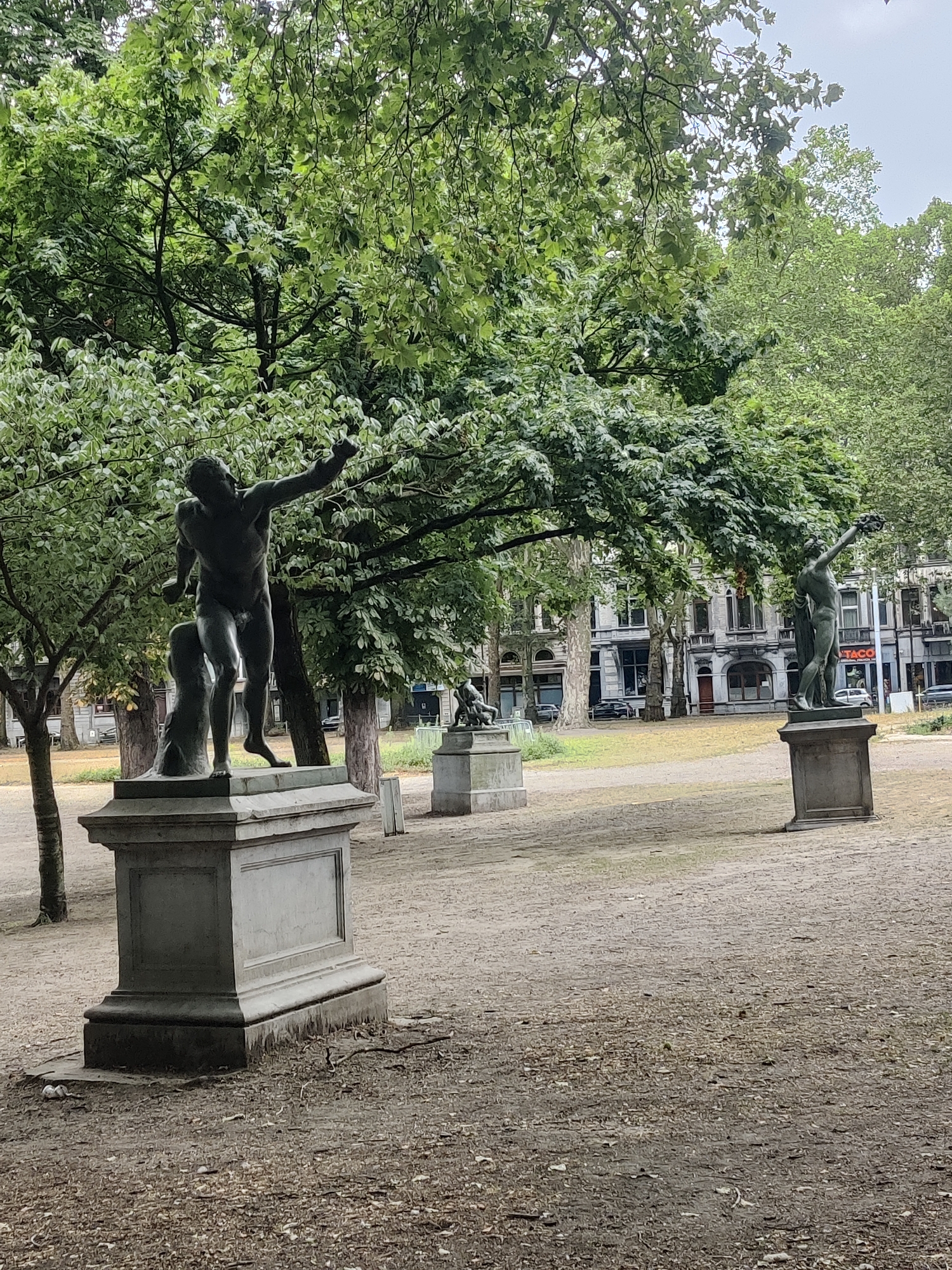 The image depicts a park setting with multiple bronze statues on pedestals, located in Liège, Belgium. In the foreground, a statue of a male figure with outstretched arm stands on a stone pedestal. Behind it, other statues are visible, some partially obscured by trees. The ground is covered with gravel and fallen leaves. Lush green trees with dense foliage surround the statues, creating a shaded atmosphere. In the background, classical-style buildings line a street, with cars parked along the sidewalk, suggesting an urban environment adjacent to the park. The sky is overcast, casting a diffused light over the scene.