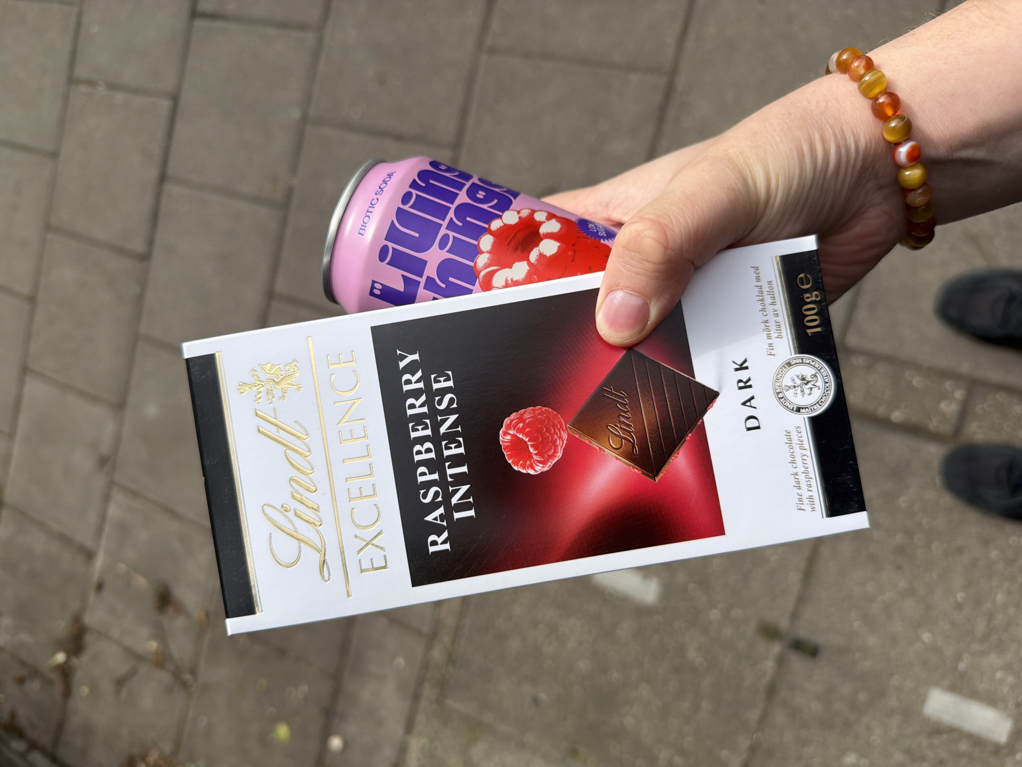 A close-up, slightly tilted shot shows a person's hand holding a Lindt Excellence Raspberry Intense Dark chocolate bar and a pink Living Things biotic soda can. The hand, adorned with a beaded bracelet featuring orange, yellow, and white stones, is positioned in the center-right of the frame. The background is a blurred outdoor setting, likely a paved sidewalk or street with grey brick patterns, and a dark shoe is partially visible at the bottom right.
The chocolate bar is prominently displayed, featuring the text "Lindt EXCELLENCE RASPBERRY INTENSE DARK" in gold and white lettering. It also states "Fine dark chocolate with raspberry pieces" and "Fin mörk choklad med bitar av hallon," indicating a Swedish translation. The weight is "100g e," and "LINDT & SPRÜNGLI MAITRE CHOCOLATIER DEPUIS 1845" is visible on a gold seal. A visual of a raspberry and a piece of chocolate is on the packaging.
Behind the chocolate bar, a metallic pink soda can is held, with "Living Things" and "BIOTIC SODA" clearly visible. A graphic of a raspberry-like jelly or fruit is on the can, along with "LOW SUGAR." The bright, natural lighting suggests it is daytime. The overall impression is a casual outdoor moment, possibly a snack or drink break in an urban environment like London.