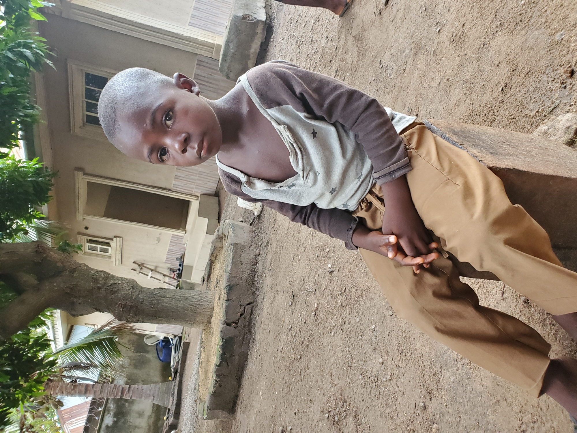 A young, dark-skinned boy with a closely shaved head sits on a stone or concrete ledge in an outdoor setting. He wears a long-sleeved shirt with brown sleeves and a lighter colored torso, possibly a t-shirt layered underneath, and khaki-colored pants. His hands are clasped in front of him. The ground is dirt. Behind him is a building with a window and a doorway visible. A tree with green foliage is present on the left side of the frame. A ladder, some water jugs, and other unidentified objects are near the building. The location is Mararaba, Nigeria.