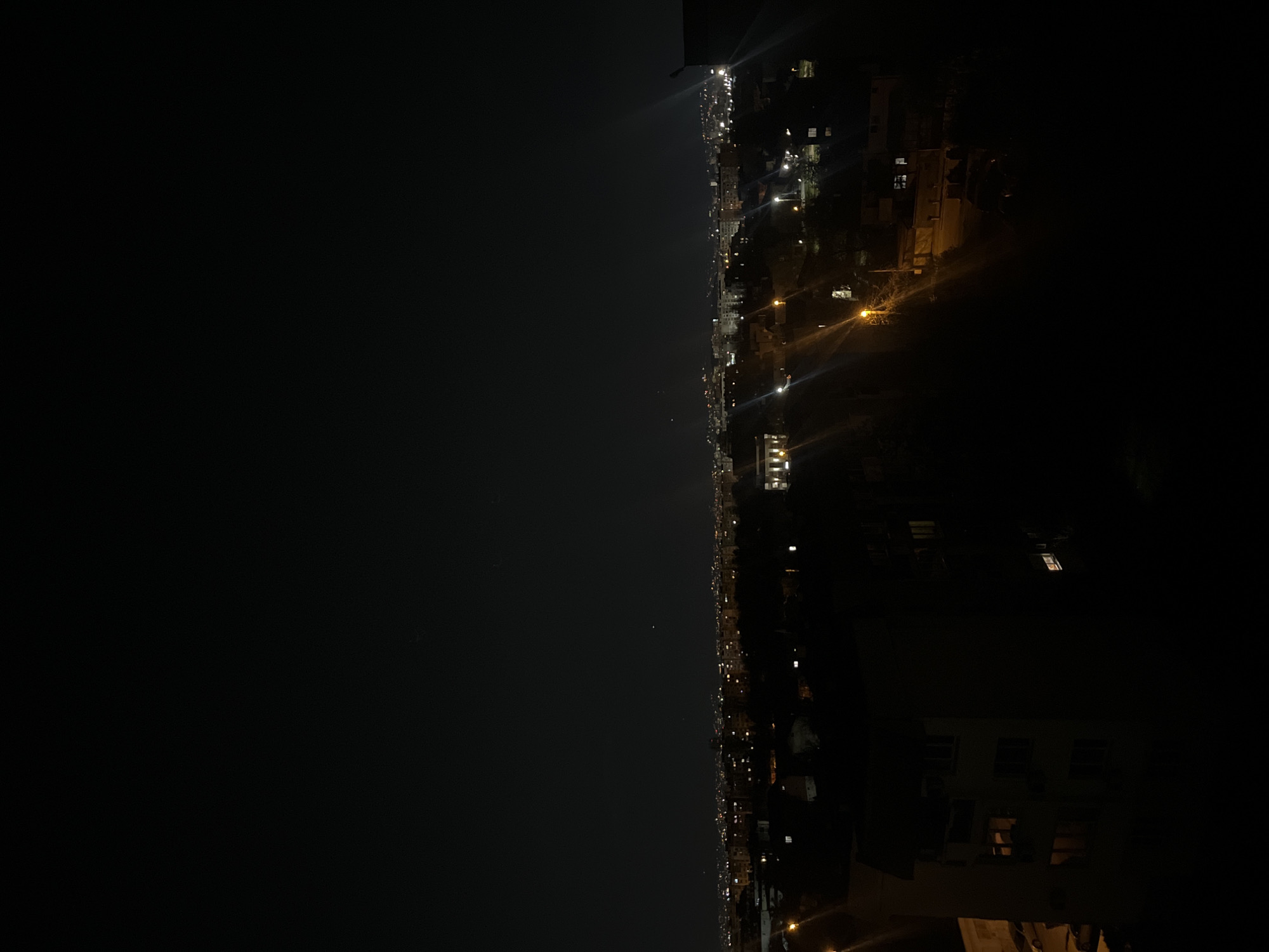 The image depicts a nighttime cityscape, likely from a high vantage point in Sofia, Bulgaria. The left portion of the frame is dominated by a dark, unilluminated sky. The right portion shows a city with streetlights, residential buildings, and commercial structures, some with illuminated windows. The scene is generally dark with points of light emanating from the city. The overall impression is of a tranquil night view of a city.