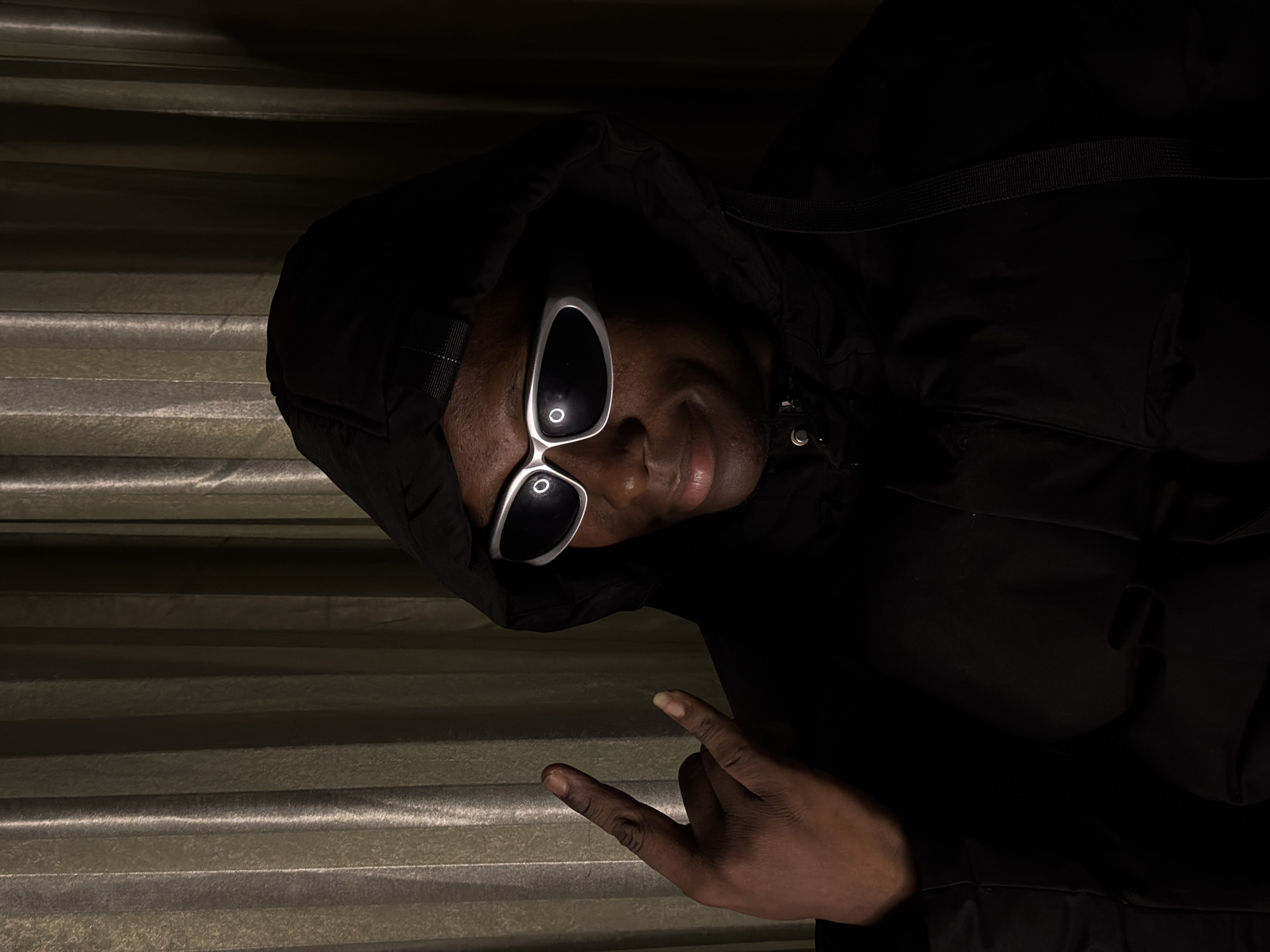 A person wearing a black hooded jacket and silver-rimmed sunglasses is shown from the chest up, with their head turned to the right and their left hand raised, forming a "rock on" gesture. They are standing in front of a backdrop of green, vertically-pleated curtains. The lighting creates strong shadows, with the left side of the person's face and body well-lit and the right side mostly in shadow. The image was taken in the city of Jiangjin, China.
