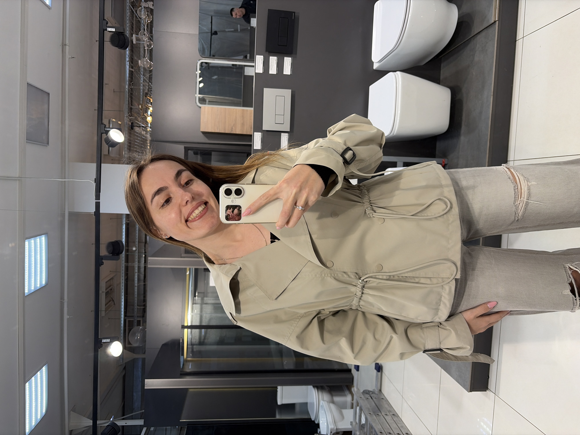A woman is taking a selfie in what appears to be a bathroom showroom. She is holding a smartphone in her right hand, with her face smiling toward the camera. The background features wall-mounted toilets and a display of bathroom fixtures. The location is identified as Izhevsk, Russia, based on the available metadata.