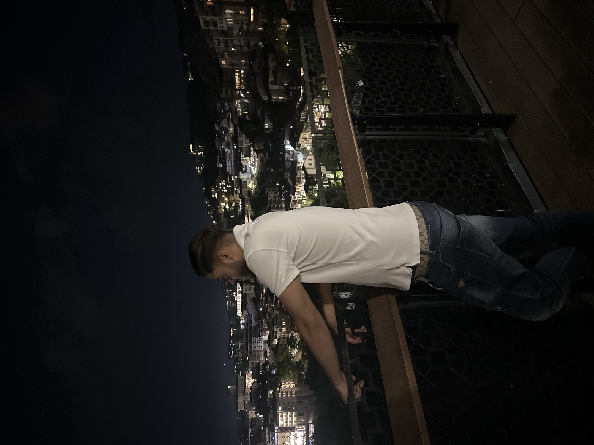 A man is standing on a balcony overlooking a city at night. The man has dark hair and a beard, and he is wearing a white t-shirt and blue jeans. He is leaning over the railing of the balcony, looking down at the city. The city is lit up with lights, and there are buildings of various heights. The sky is dark. The location appears to be high above the city.