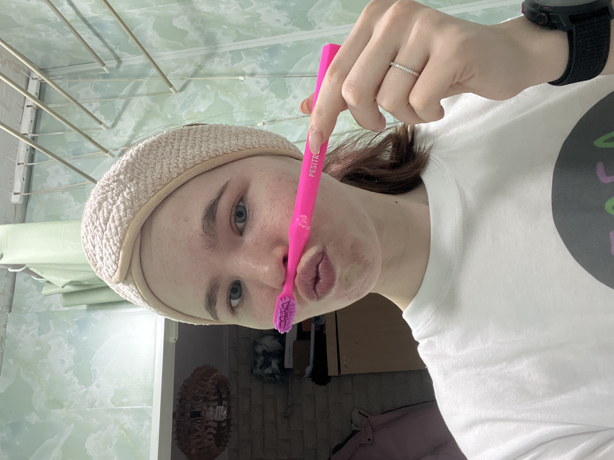 A person is posing with a pink toothbrush held vertically in front of their face, the brush end touching their lips. The person has fair skin, blue eyes, and is wearing a beige headband and a white t-shirt with a design on it. A ring is visible on their finger and a black smartwatch is on their wrist. The background shows a room with a light green and white patterned wall, laundry drying on a rack, and glimpses of furniture and decor. The location is identifiable as Chaikovsky, Russia, based on textual information on the toothbrush and contextual clues within the image.