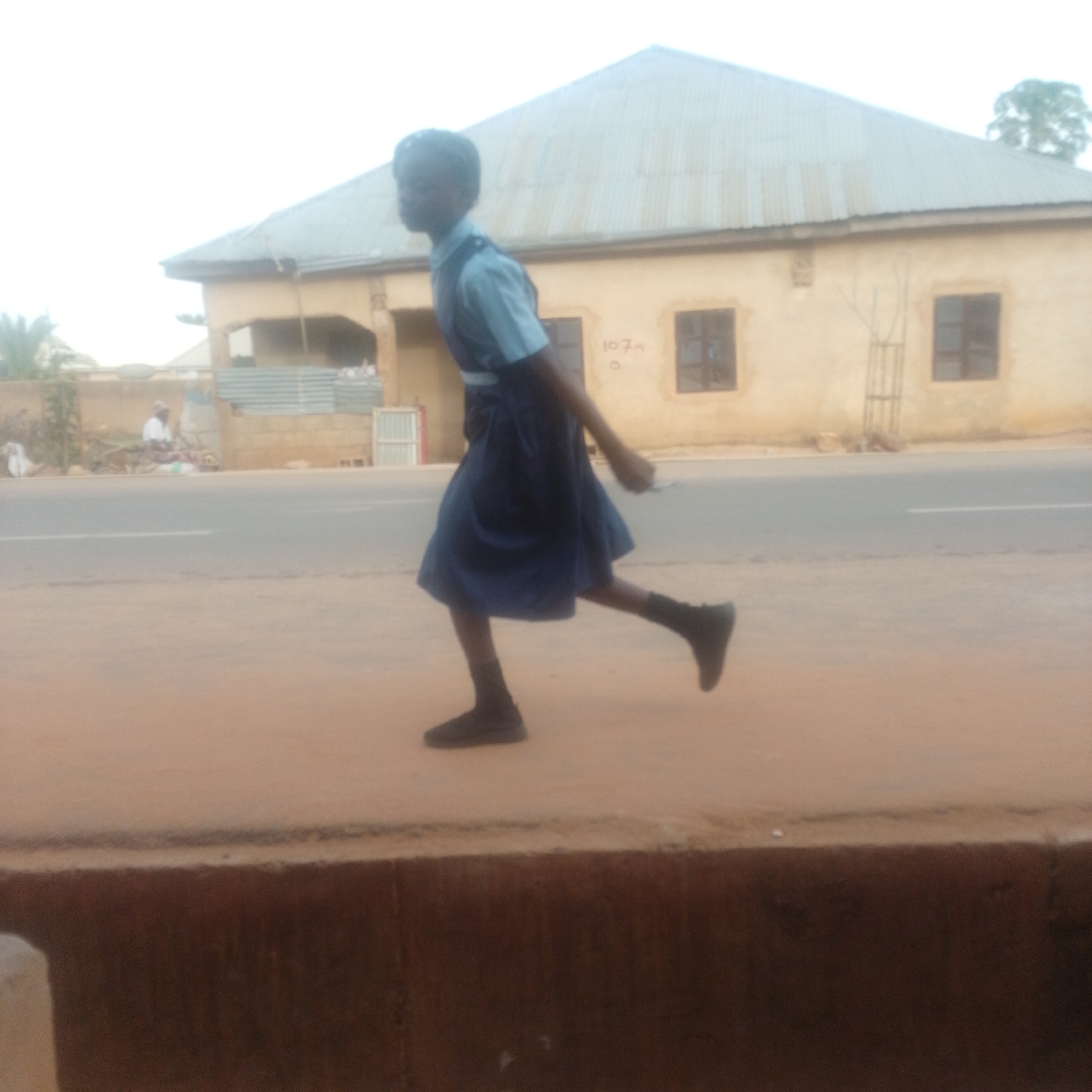 The image shows a young person in a school uniform, a blue collared shirt and a dark blue skirt, walking or running on a dirt road. The person is wearing dark shoes and dark socks. In the background, there is a light-colored building with a corrugated metal roof and windows. A road is visible in front of the building, with a white line marking the center. To the left of the building, there are some trees and what appears to be a fence made of corrugated metal. Further to the left, a person in a white shirt is sitting near some goods, possibly at a market stall. The sky is overcast. The scene appears to be in an urban or semi-urban setting, possibly in Gombe, Nigeria, given the architectural style of the building and the general environment. The overall lighting suggests it is daytime.