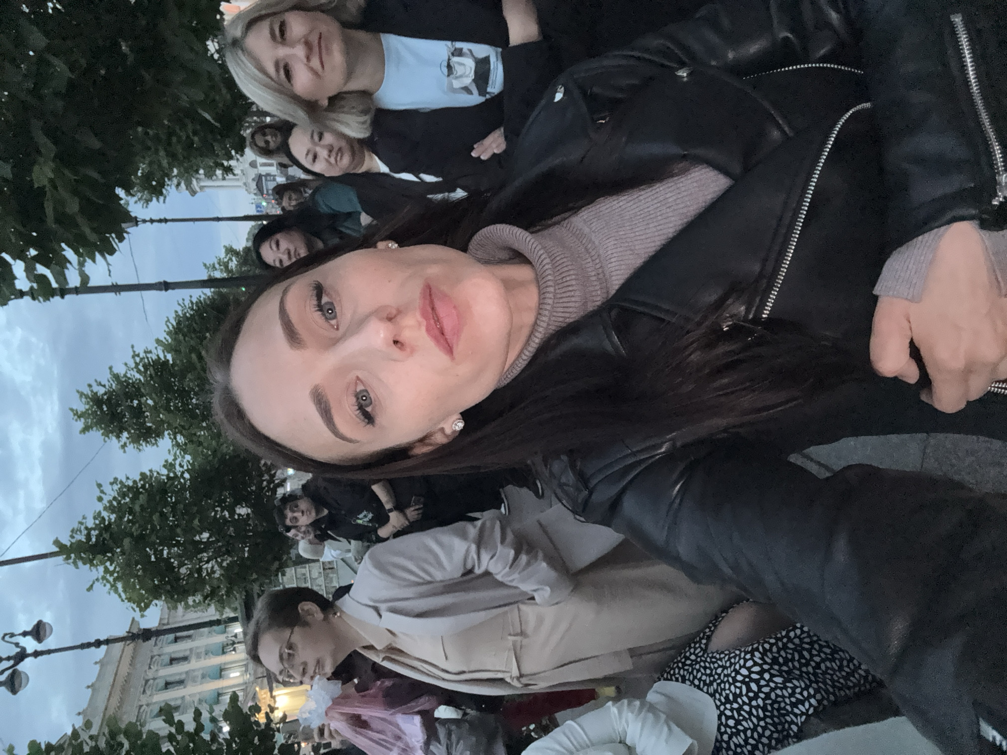 This is a selfie taken outdoors in Saint Petersburg, Russia, during the evening. The foreground is dominated by the face and upper body of a woman with dark hair, wearing a black leather jacket over a light brown turtleneck sweater. She is looking directly at the camera with a neutral expression.
Behind her, a crowd of people are visible, with a mix of men and women. To the left, a woman wearing glasses and a light-colored suit is looking to her left. Further back, other individuals are looking in various directions. The background shows trees and classical architecture, consistent with the cityscape of Saint Petersburg, including a lamppost and buildings with ornate facades. The sky is overcast and appears to be twilight. The overall atmosphere suggests a gathering or event taking place in a public urban space.