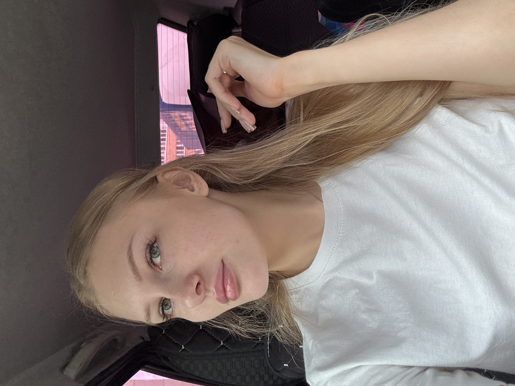 A young Caucasian woman with long blonde hair and blue eyes is pictured from the chest up, inside a vehicle. She is wearing a white t-shirt and has a light-colored necklace. Her right hand is raised to her face, with her fingers gently touching her cheek. The background shows the interior of a car, with a dark-colored seat and a pinkish-tinted window reflecting buildings. The lighting suggests it is daytime. The specific location in the city of Perm, Russia cannot be determined from the visual information present.