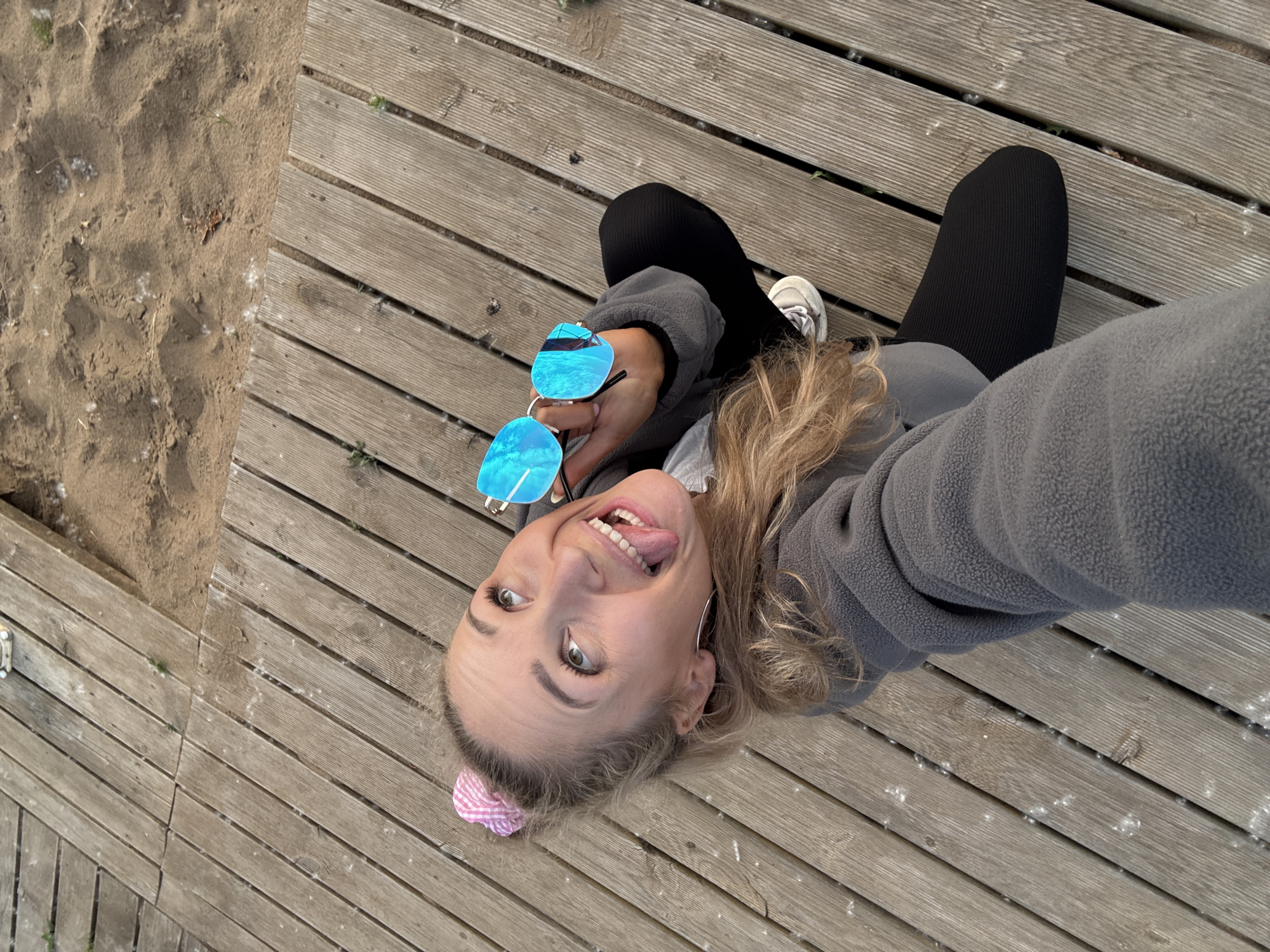A female individual is depicted in a high-angle selfie photograph taken outdoors in Chaikovsky, Russia. She is a light-skinned individual with blonde hair tied in a half-up style, secured with a pink and white checkered scrunchie. She wears large silver hoop earrings. Her face displays an expressive smile with an extended tongue, and her eyes are directed upwards and to the viewer's left. She is dressed in a grey fleece pullover, black ribbed leggings, and white sneakers with dark laces on her visible left foot. Her right hand holds a pair of blue mirrored sunglasses with a silver frame. Her posture suggests she is kneeling or squatting on a wooden surface. The setting is a wooden boardwalk constructed from light brown, parallel planks, which shows signs of wear and minor white, fluffy debris (possibly pollen or seeds). Adjacent to the boardwalk, a section of light brown sand is visible, also strewn with similar white debris and sparse green vegetation.