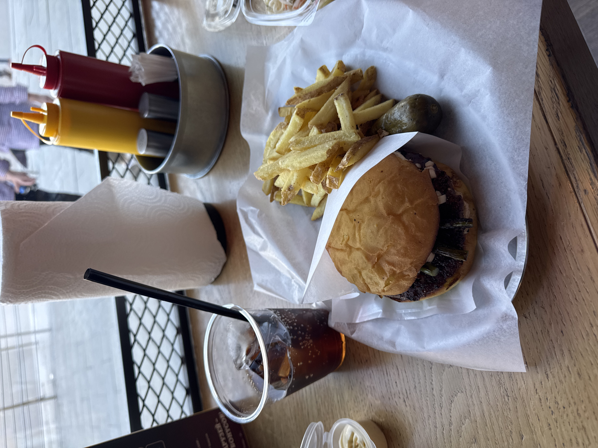 This is an overhead shot taken on a wooden table. There is a burger in a white paper tray, next to a pile of fries and a pickle. A metal container holds ketchup, mustard, and utensils. A cup of soda with a black straw is also on the table, along with a napkin and small containers. There is metal rack in the background. The location is likely a restaurant. There is no evidence to suggest this is Moscow, Russia.