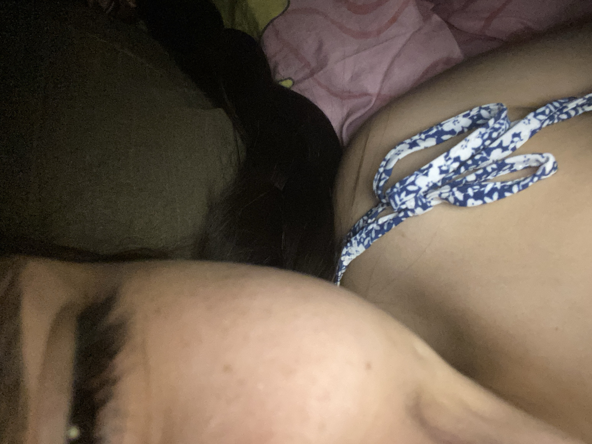 This close-up image shows a person in a dimly lit, indoor setting, likely resting on a bed or couch. The upper left portion of the image features the side of a person's head, revealing their forehead, eyebrow, and part of an eye. Long, dark hair cascades down the left side, partially obscuring the face.
To the right of the hair, a body part, possibly a shoulder or torso, is visible, fair-skinned and smooth. A blue and white patterned strap, consistent with swimwear or lingerie, crosses this area. Further to the right and above, a pink fabric object, possibly a blanket or pillow, is partially visible with some lighter, faded patterns. The overall lighting is low, suggesting it might be nighttime or a darkened room. No specific activity is clearly depicted other than the person appearing to be at rest. No text is visible in the image. The image lacks specific location cues for Cotoperíz, Venezuela.