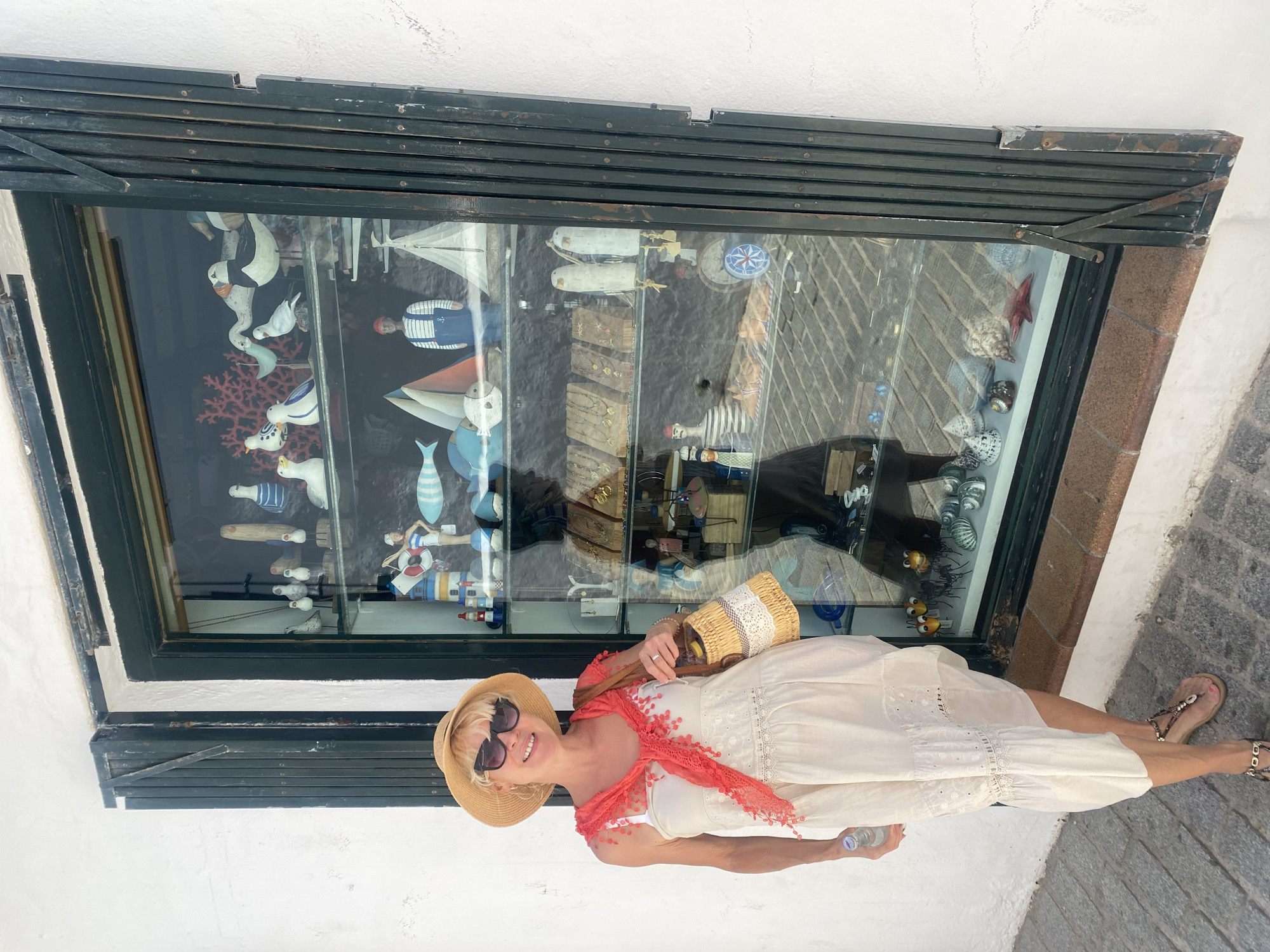 A woman is standing outside a shop in Conil De La Frontera, Spain, posing for a photograph in front of a shop window filled with nautical-themed decorations. The woman, who has blonde hair and is wearing sunglasses and a straw hat, is dressed in a light-colored, sleeveless dress and a red scarf draped around her shoulders. She is carrying a woven handbag. The shop window displays a variety of decorative items, including wooden seagulls, sailboats, a sailor figurine, coral, and shells. The shop has a dark, collapsible metal security gate that is partially pulled back, revealing the glass window. The ground in front of the shop is paved with cobblestones, and the building is painted white.