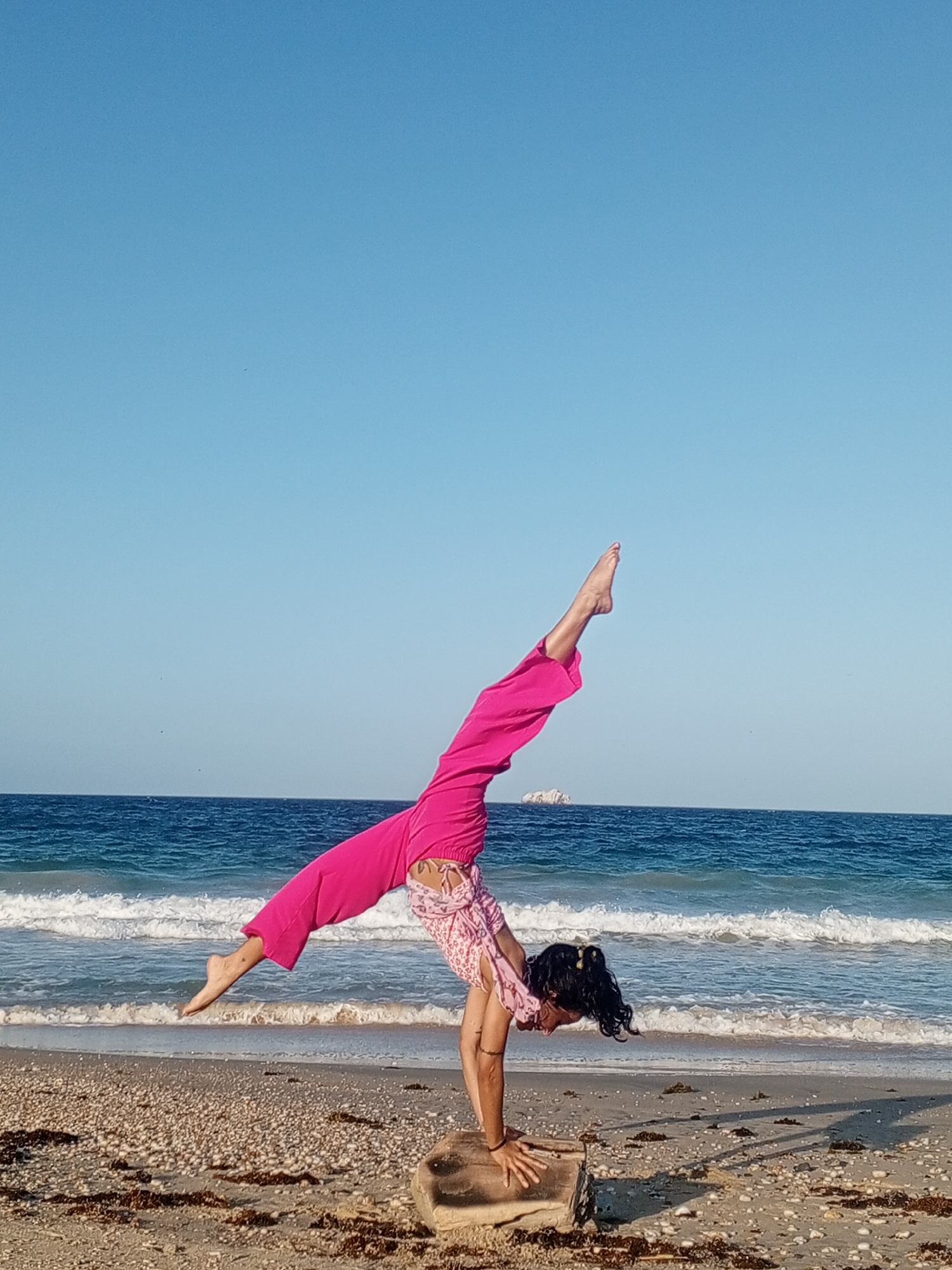A woman is performing a handstand on a beach in Porlamar, Venezuela. She is wearing a pink patterned top and pink pants. Her hands are placed on a piece of driftwood. Her legs are extended and slightly bent. The beach is sandy, with some seaweed scattered around. The ocean is in the background, with waves breaking on the shore. The sky is clear and blue. In the background, a small island can be seen in the distance.