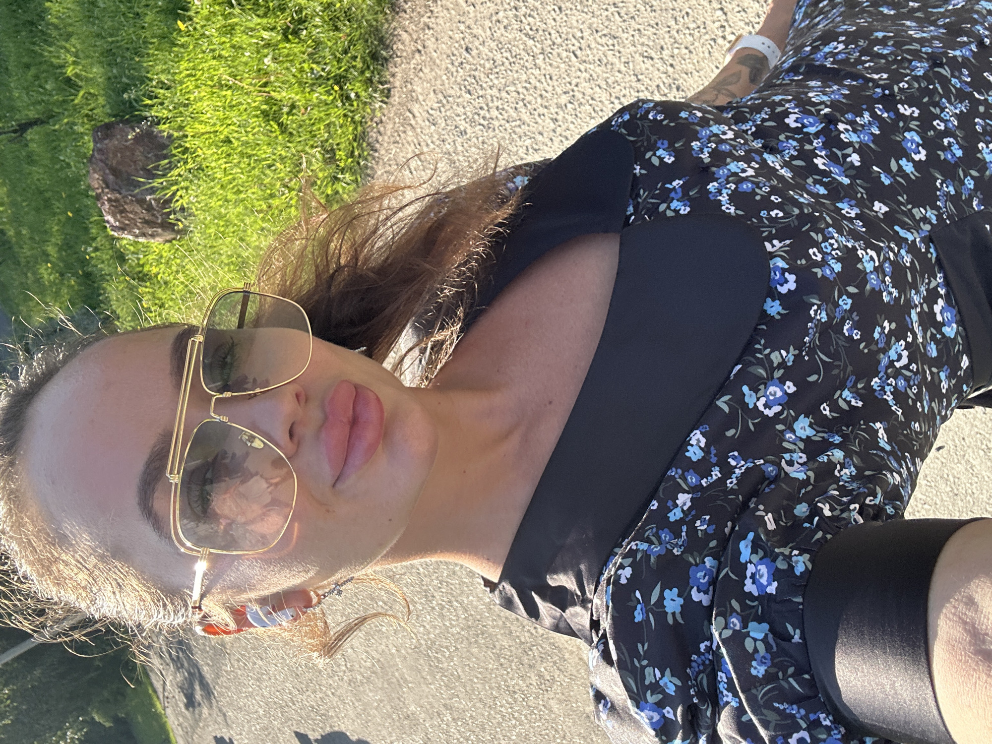 The image is a selfie taken outdoors, with a woman in the foreground. She has long, wavy brown hair and is wearing large, gold-rimmed sunglasses and a black dress with a blue floral pattern. She is also wearing silver hoop earrings and earbuds. Her lips are full and appear to be coated with lip gloss. The background shows a paved surface, green grass, and a large rock. The lighting suggests it is daytime, possibly late afternoon due to the angle of the sun. The overall setting appears to be a park or outdoor recreational area. There is no specific indication of the location being Duvankul', Russia.