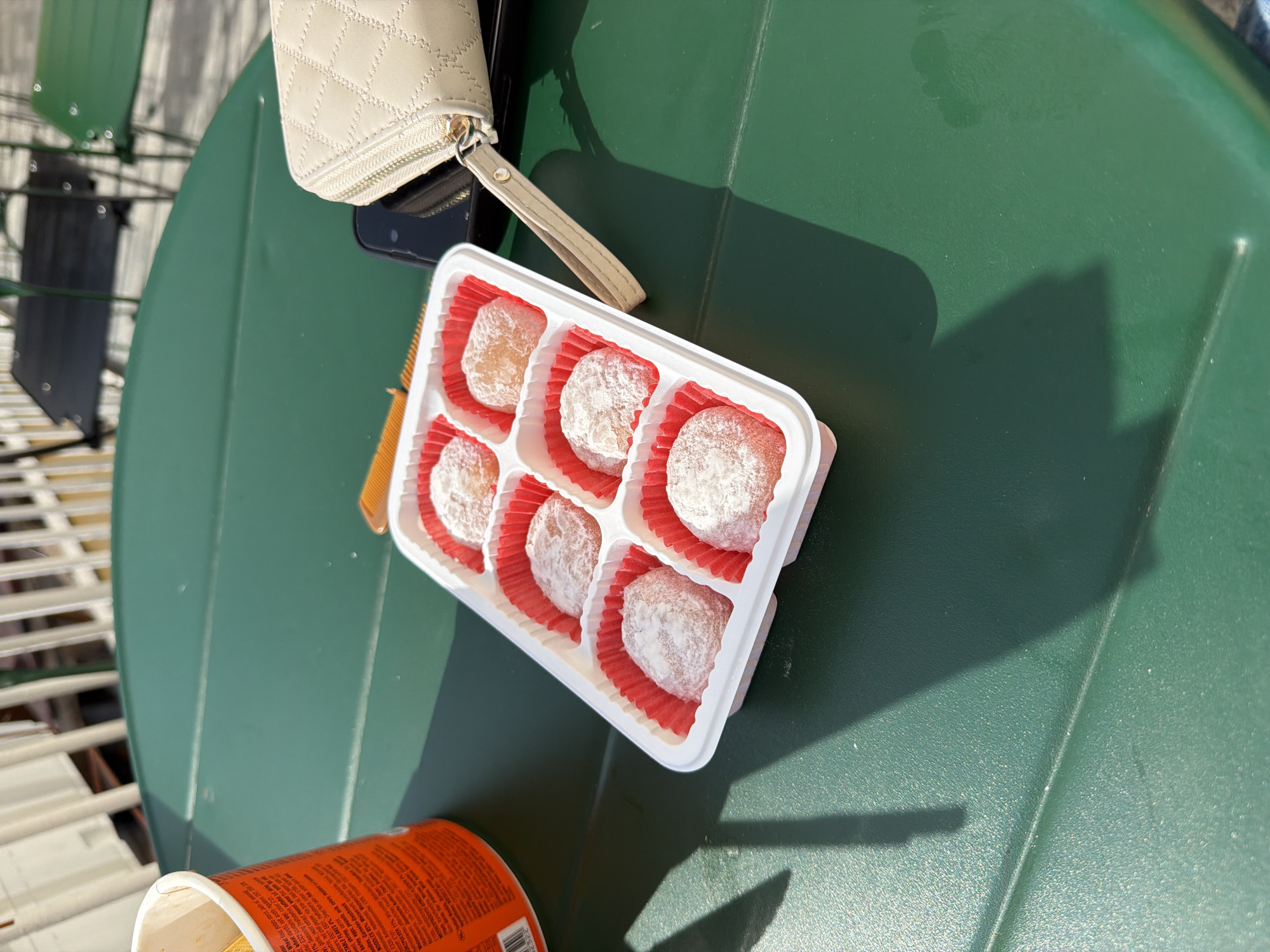 The scene depicts an outdoor setting, likely a cafe or patio, in Veliko Tarnovo, Bulgaria, under bright sunlight. A dark green, rectangular metal table occupies the foreground and midground. On the table, a white plastic tray containing six individual portions of light-colored, powdered mochi-like sweets, each in a red paper cup liner, is prominently positioned slightly off-center to the right. To the left of the tray, an orange cylindrical paper cup or container with visible English text on its side is present. To the right of the tray, a beige, quilted wallet or small pouch with a zipper and a wrist strap rests on top of a dark-colored smartphone. Shadows from an unseen overhead structure or foliage are cast across the table's surface. In the background, out of focus, white vertical railings and dark metal outdoor chairs with green seats are visible. No people are present in the immediate view, and there are no direct actions or interactions depicted.