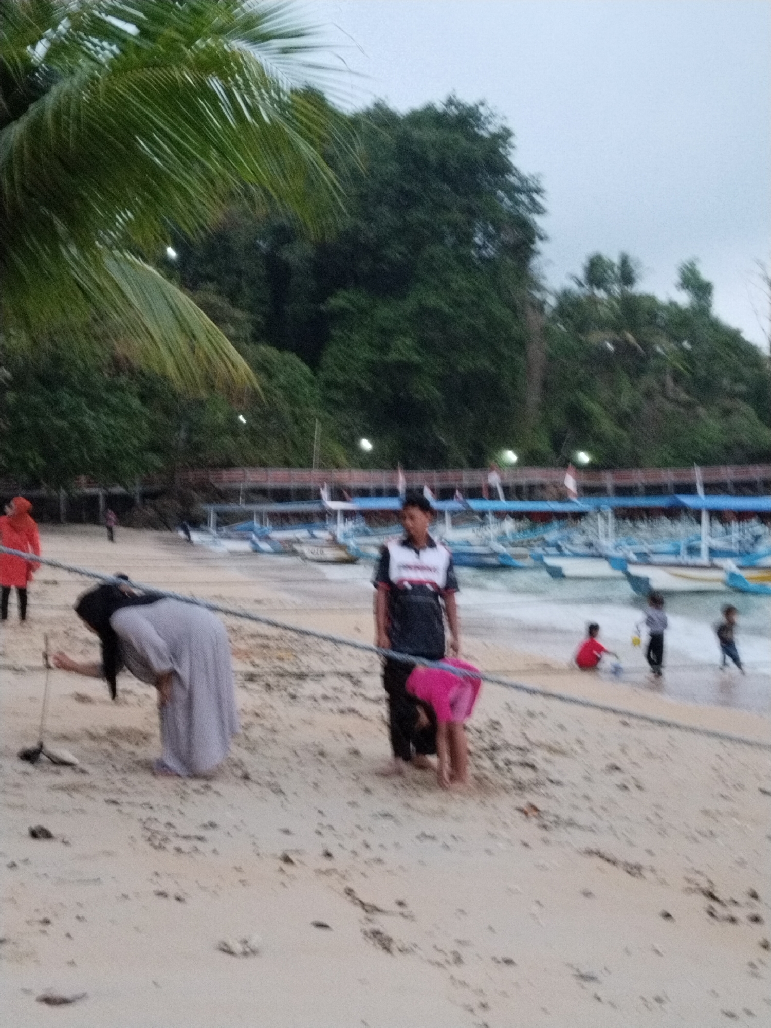 A beach scene in Prigi, Indonesia, features several individuals on a sandy shore. In the foreground, a person wearing a light gray dress and a headscarf is bent over, sweeping the sand with a broom. To their right, a person in a dark shirt with a white and red collar stands looking towards the left. A child in a bright pink garment is on their hands and knees in front of this person. Further back, a person in a red coat stands on the beach. In the background, multiple boats are docked in the water, and a line of trees forms the treeline. The sky is overcast.