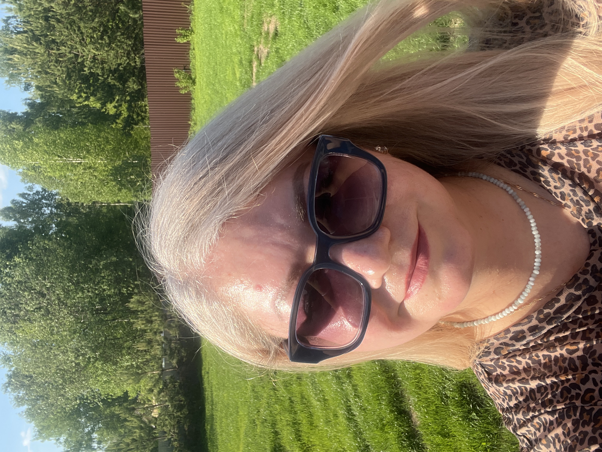 A woman with blonde hair, wearing sunglasses and a leopard-print top, is posing for a photo outdoors. She is also wearing a beaded necklace and an earring is visible on her left ear. The background shows a green lawn, trees, and a brown fence. The photo appears to be taken in the daytime, likely in Новая Чернушка, Russia.
