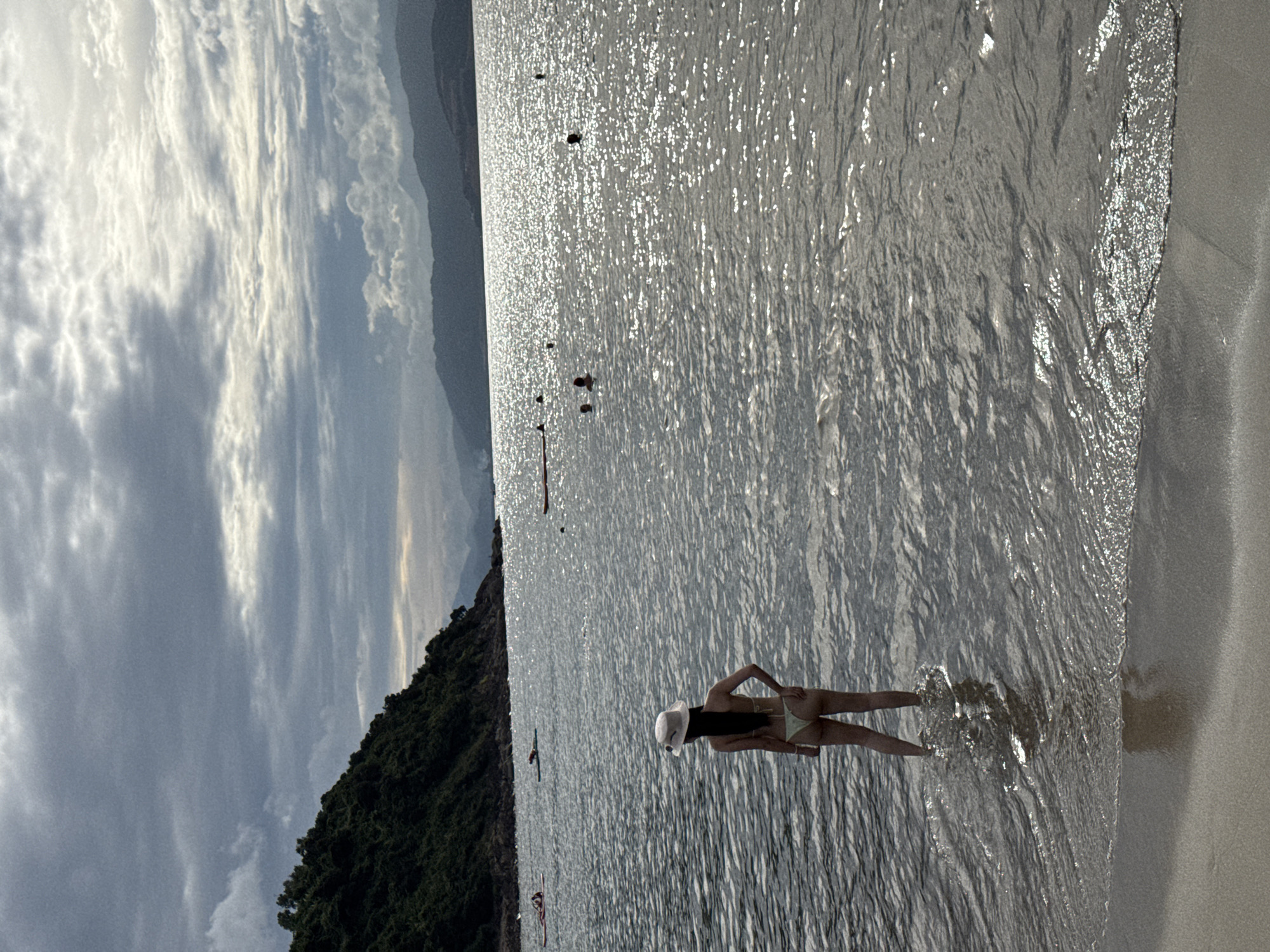 This image depicts a serene beach scene at sunset, likely on a coastline near Da Nang, Vietnam, characterized by a dramatic, cloudy sky and the shimmering surface of the sea. In the foreground, a woman stands waist-deep in the calm, shallow water, wearing a light-colored bikini and a white sun hat. Her back is to the viewer as she gazes out at the expanse of the ocean. The water is a dark, reflective gray, rippling gently with the incoming tide, and the sandy shore is visible in the bottom left corner.
Further out in the water, several other individuals are visible, some swimming or wading, and a few are on paddleboards. Two paddleboarders are positioned further back, closer to a verdant, tree-covered hillside that slopes down to the water on the left side of the frame. The background features a range of distant, hazy mountains silhouetted against the diffused sunlight breaking through the clouds. The overall atmosphere is one of tranquility and natural beauty, with the setting sun casting a soft, atmospheric glow over the landscape.