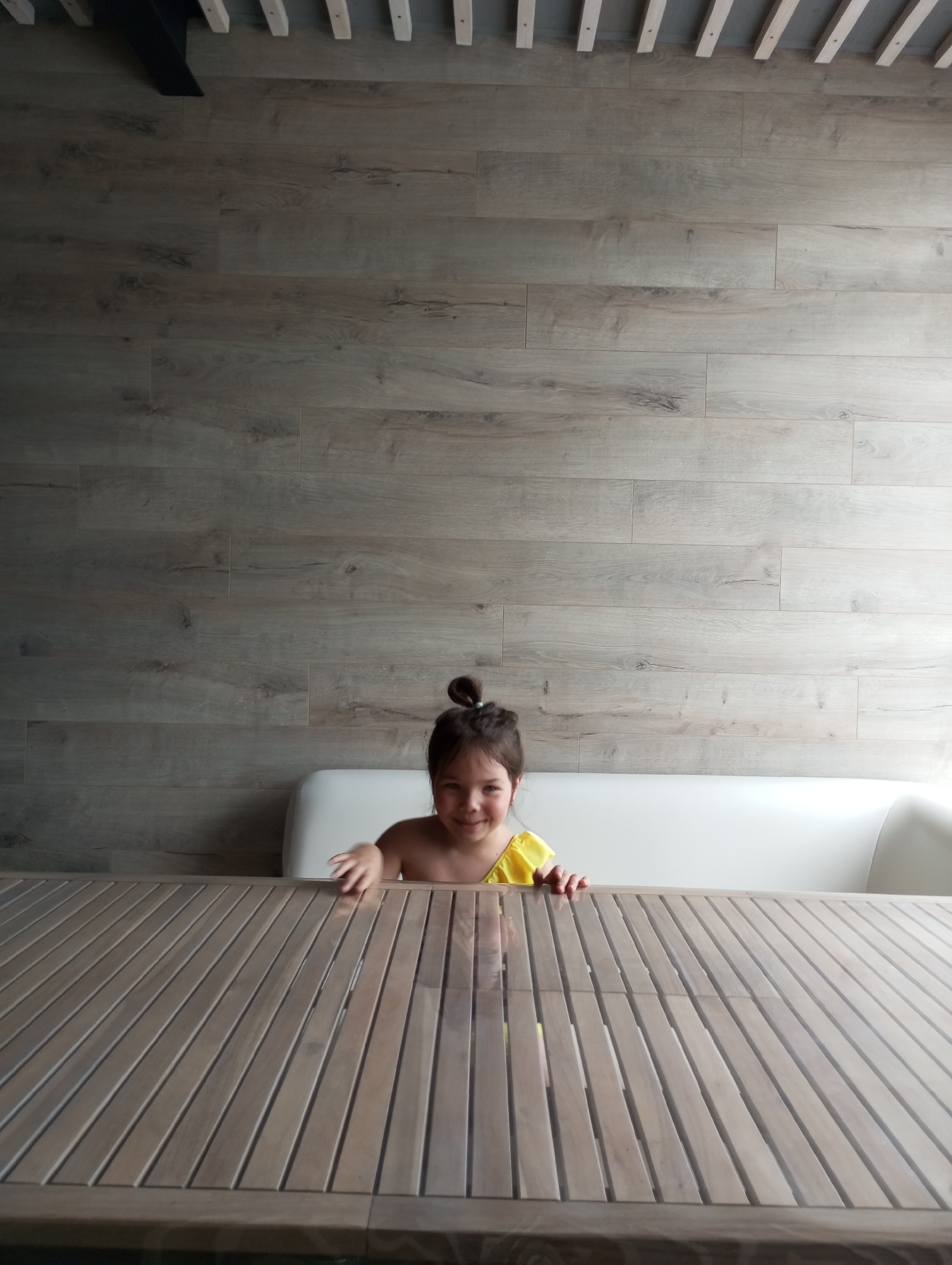 A young girl in a yellow top is sitting at a wooden table in front of a white couch. The wall behind her is light brown with a horizontal plank pattern, and there are wooden beams on the ceiling above. The location and city cannot be determined from the given information.