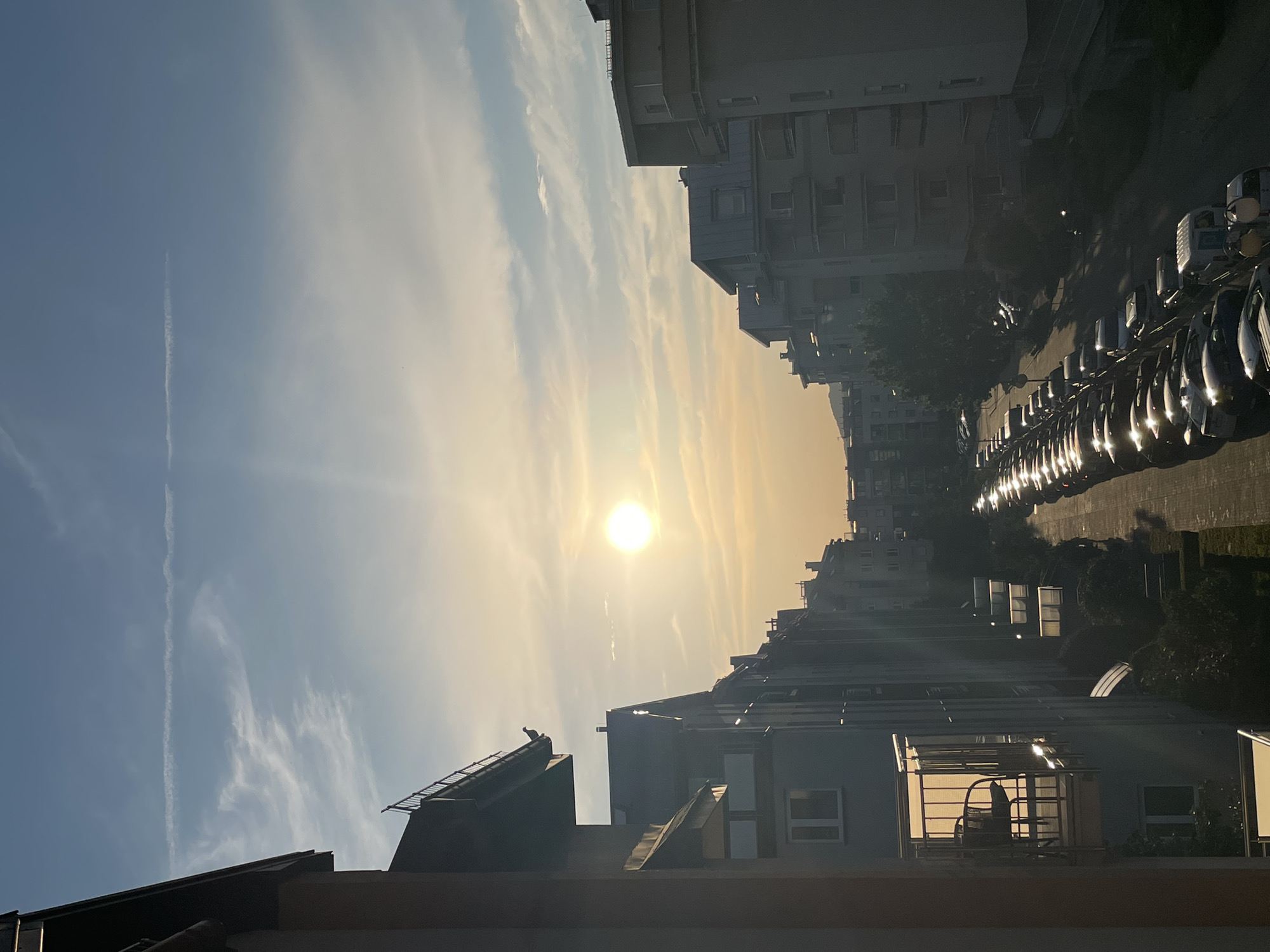 The image depicts an urban scene at sunset, with the sun shining brightly in the sky, casting a warm, golden glow. The sky is partially cloudy, with wispy cirrus clouds and a prominent contrail. In the foreground, several rooftops of apartment buildings are visible. A lone bird is perched on the edge of one of the rooftops.
The middle ground features a row of parked cars, their headlights reflecting the setting sun. Beyond the cars, more apartment buildings line a street, their facades illuminated by the warm light. The buildings appear to be modern, with balconies and multiple windows. The overall impression is one of a quiet evening in a residential urban area. The time of day is clearly sunset, indicated by the low angle and warm color of the sun, as well as the long shadows cast by the cars and buildings. The weather appears clear, with only some clouds present. There are no discernible location cues that would indicate a specific city or region. No text is visible in the image.