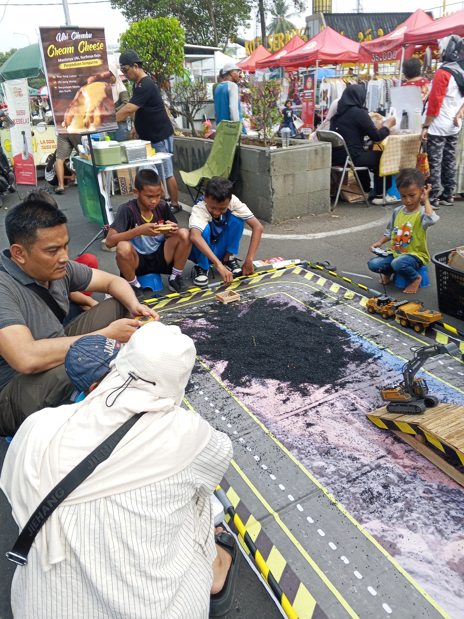 The image captures an outdoor scene in Serang, Indonesia, possibly during a market or festival. In the foreground, several individuals are gathered around a large, printed mat depicting a road with a construction site. A man in a gray polo shirt is seated, examining a small object in his hand, while a child in a blue cap is seated nearby. Another person, wearing a white hooded garment and a striped shirt, is also seated, with a black strap with the word "JIEHANG" visible across their back.
In the middle ground, two young boys are playing with toy construction vehicles on the road mat. One boy in a yellow shirt is holding a remote control, actively engaged with a toy excavator. The other boy, in a blue shirt and black shorts, is also playing with the toys. Behind them, a table displays food items, including a poster advertising "Ubi Gilembu Cream Cheese." Several people are milling around this area, including a man interacting with a woman behind the table.
The background shows various stalls and tents, some adorned with red canopies. Merchandise, possibly clothing, is displayed on racks. The environment suggests a lively public gathering with commercial activities. The presence of a poster in Indonesian, along with the general atmosphere, points to this location being in Indonesia, specifically Serang as indicated by external information. The overall scene depicts a moment of recreation and commerce in an urban outdoor setting.