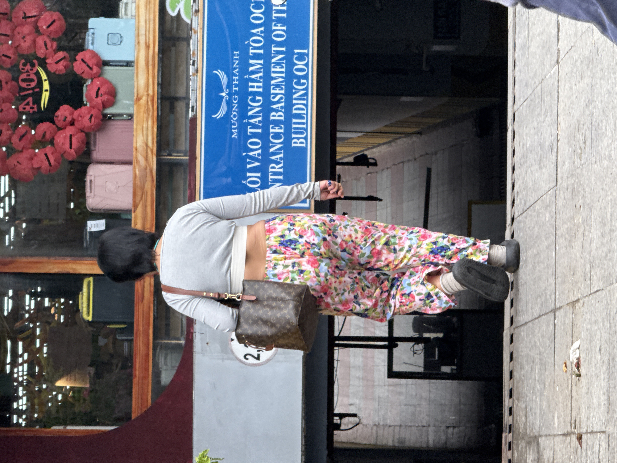 A woman is walking away from the camera in an outdoor urban environment. She has short black hair and is wearing a light gray cropped long-sleeved shirt, colorful floral patterned pants, white socks, and dark slip-on shoes. She is carrying a brown Louis Vuitton-patterned bag over her left shoulder. Behind her, a blue sign is visible with text in Vietnamese and English that reads "MƯỜNG THANH ENTRANCE BASEMENT OF TH BUILDING OC1". The sign is mounted above an entrance that leads down into a dark area. To the left of the sign, a circular sign with the number "2" is visible. The woman is walking on a paved walkway. In the background, there is a storefront window displaying various luggage items, including pink, green, and light blue suitcases, and a cluster of red decorations. The overall setting appears to be a street in Nha Trang, Vietnam.