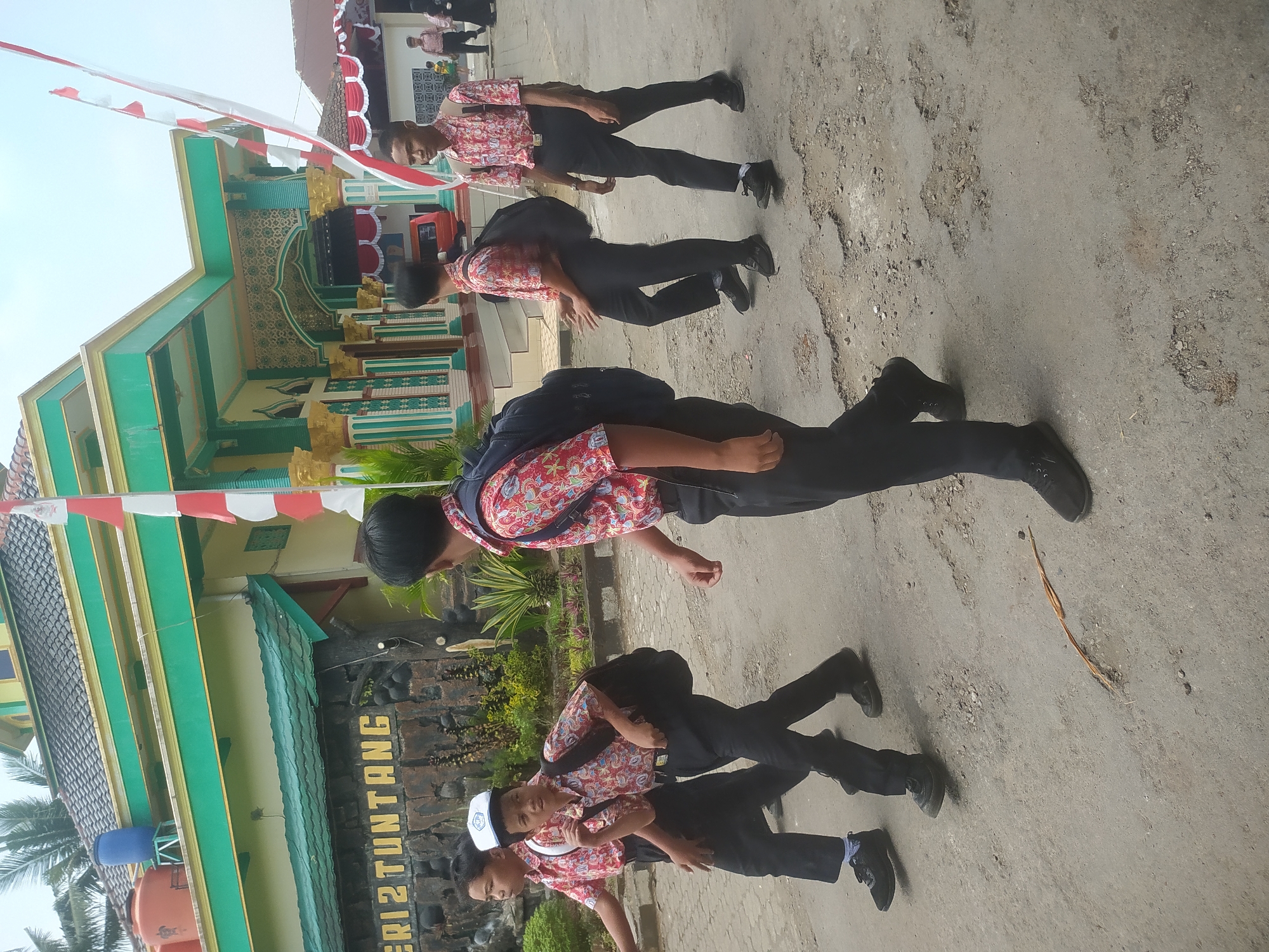 In Candirejo, Indonesia, a group of young male students in uniform are walking on a paved road in front of a building. The building has green and yellow accents and features Indonesian flags. The students are wearing collared shirts with a floral pattern, black pants, and dark shoes. Some of them have backpacks. The road has visible potholes and debris. A sign with the words "ERI 2 TUNTANG" is visible on the building.
