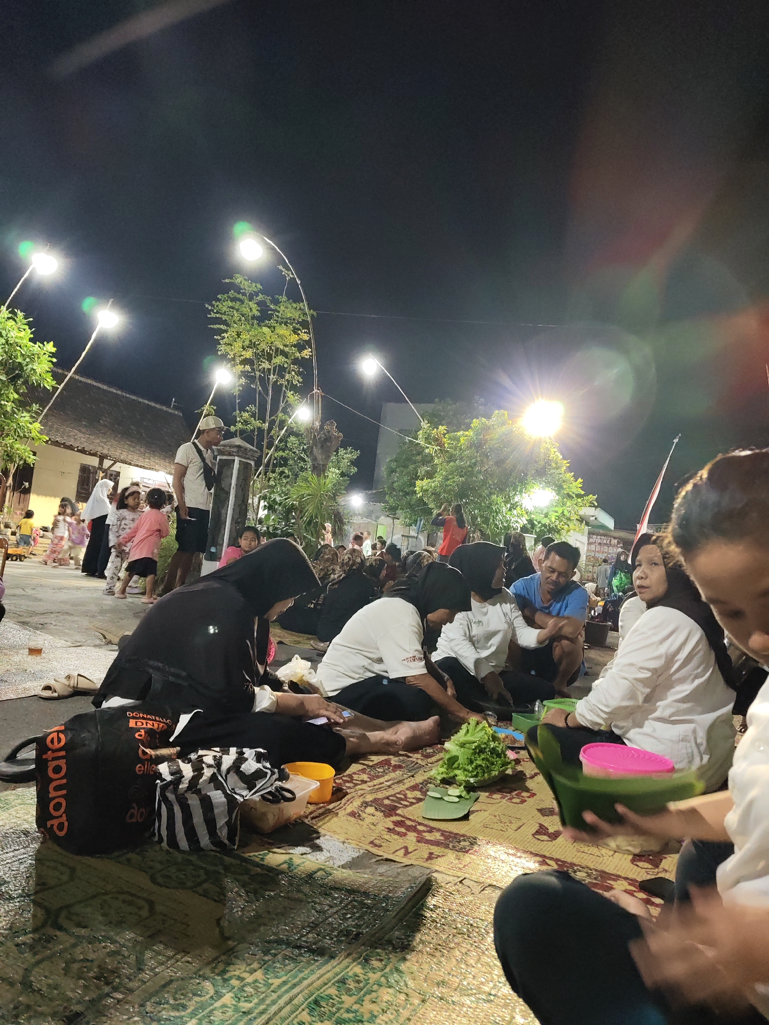 The image depicts an outdoor gathering at night in Gumpang, Indonesia. A group of people, including adults and children, are seated on woven mats on the ground. They appear to be eating or socializing. Some individuals are wearing traditional Indonesian attire, such as headscarves and long skirts. The scene is illuminated by several streetlights, which cast a warm glow on the surroundings. In the background, there are buildings and trees, suggesting a semi-urban or rural setting. Several objects are scattered on the mats, including food containers, a bag, and a striped bag. The overall atmosphere appears relaxed and communal.
