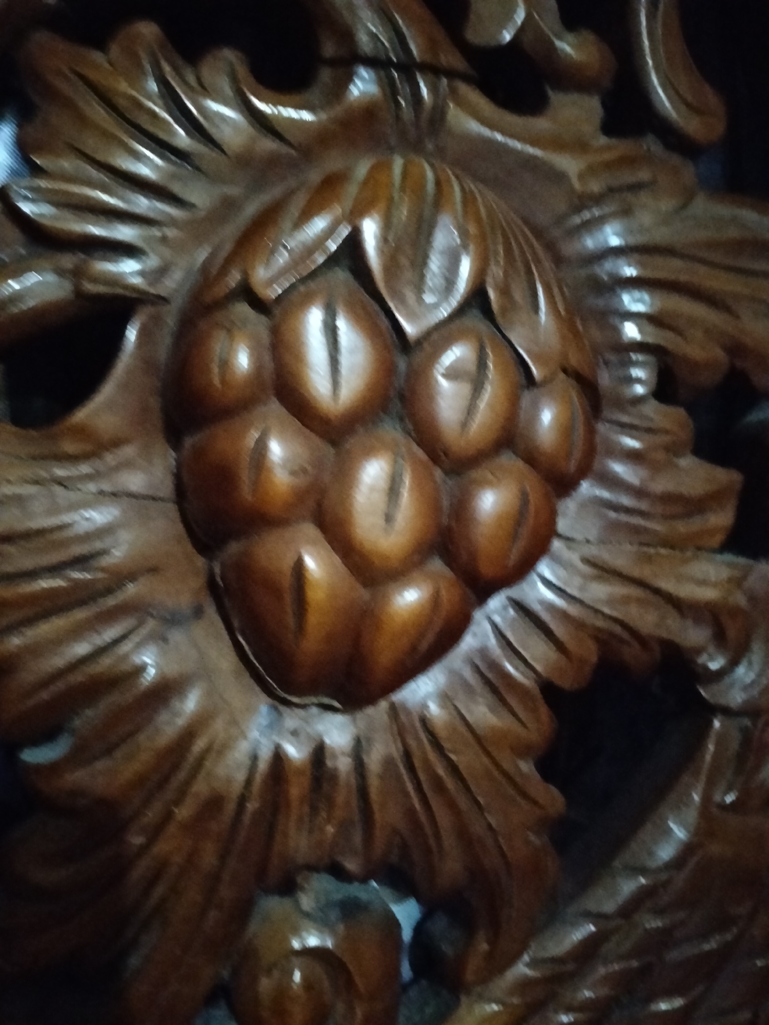 This is a close-up, low-angle photograph of a wooden carving. The carving depicts a cluster of rounded, segmented elements, resembling seeds or fruit, at its center. These are surrounded by intricately carved, petal-like or leaf-like shapes that radiate outwards. The wood has a rich, polished, reddish-brown hue, with highlights and shadows that emphasize the depth and texture of the carving. The background is dark and out of focus, suggesting the carving is part of a larger object or installation.
The image does not contain any information about the city of Payagaboh, Indonesia, nor does it depict people, actions, or interactions. The description is limited to the visual details of the carved object.
