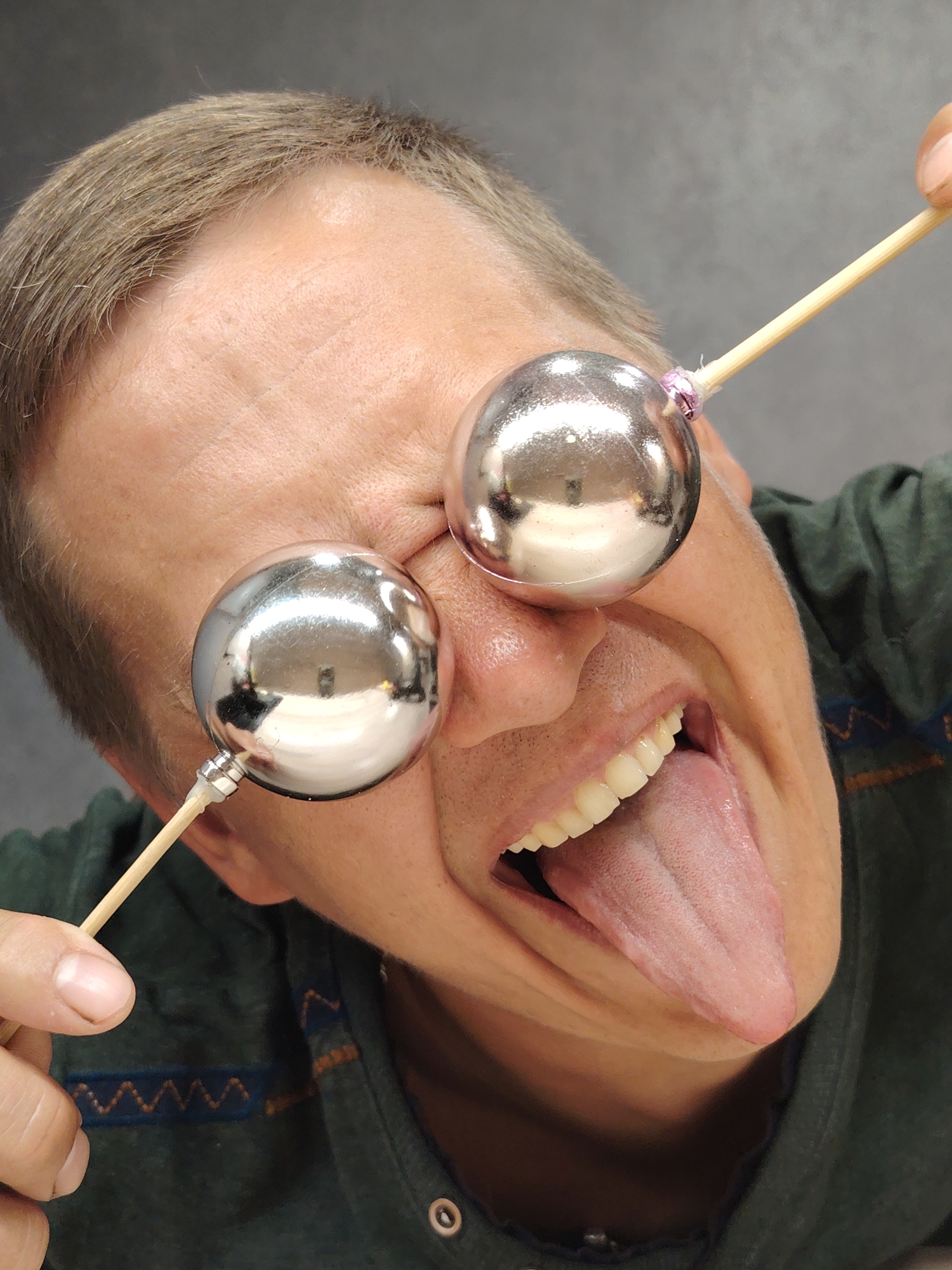 This is a close-up, eye-level shot of a person's face from the nose up, viewed from a slightly upward angle. The person, with light brown hair and fair skin, is holding two silver, spherical ornaments on wooden skewers in front of their eyes, obscuring them. Their mouth is open wide, revealing teeth and a long, pink tongue that is sticking out. The person is wearing a dark green shirt with a zigzag pattern on the collar area. The background is a plain, gray wall. The image does not contain any information that would indicate its location in Chaikovsky, Russia.