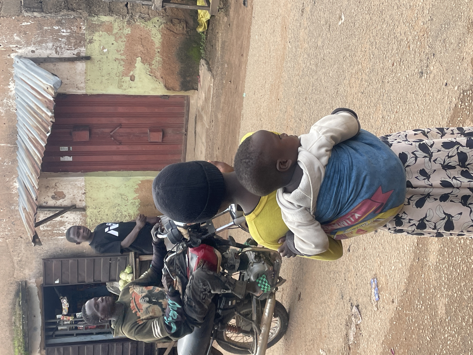 In Jos, Nigeria, a man sits on a motorcycle, his left hand on the handlebars, and appears to be looking off to the side. He wears a dark green hooded sweatshirt with a graphic design on the front and black pants. In front of the motorcycle, a child wearing a black beanie and a yellow shirt is visible from the back, with a baby wrapped in a blue and red cloth carrier on their back. The baby wears a white hooded sweatshirt. The child and baby are facing away from the viewer. Behind the man on the motorcycle, another young person stands in front of a building with a red door and a weathered corrugated metal roof. The building is painted in shades of green and brown, with peeling paint and rough textures. The ground is a dusty, gravelly surface. The overall scene depicts a moment of everyday life in an urban setting in Jos.