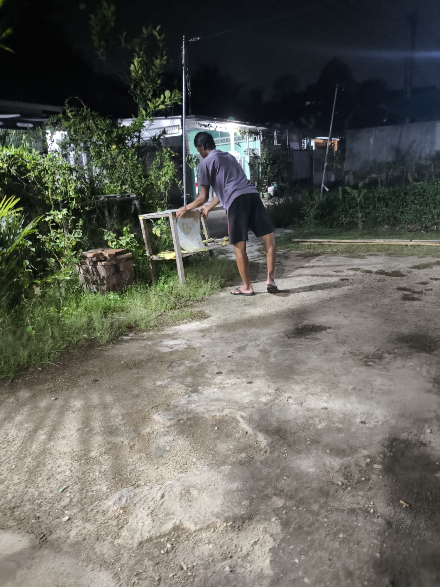 A full-body shot shows a young adult male, dressed in a purple t-shirt and black shorts, standing on a dirt and concrete surface at night. He is facing away from the camera, leaning over a wooden frame with a white, translucent sheet of plastic covering it. The man's left hand is on the frame, and his right hand appears to be reaching for the plastic. He is wearing flip-flops. The environment suggests an outdoor, possibly residential area, with overgrown greenery and a structure in the background. A stack of bricks is visible to the left of the wooden frame. The lighting indicates it is nighttime, with artificial light sources casting shadows and illuminating parts of the scene. The overall atmosphere is utilitarian and somewhat unkempt.