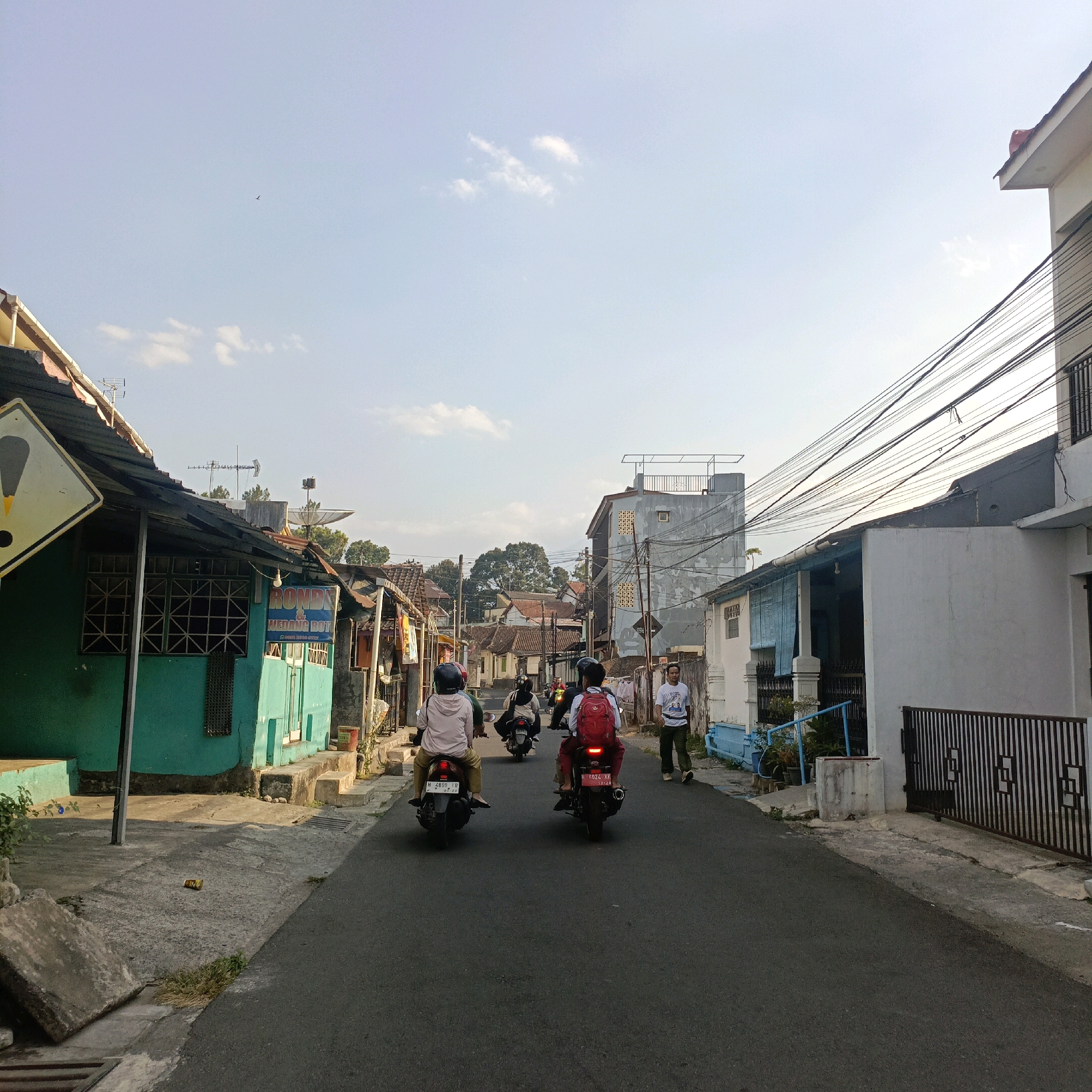 The image depicts a street scene in Salatiga, Indonesia, under a clear sky with scattered clouds. The perspective is from the rear of two motorcycles driving down a paved road. The motorcycle in the foreground has two occupants, a driver and a passenger wearing a red backpack. The second motorcycle also has two occupants. Several other motorcycles are visible further down the street, some with passengers.
On the left side of the road, there are several buildings with a weathered appearance. The closest building is painted teal and has a corrugated metal roof. A yellow diamond-shaped road sign with a black exclamation mark is affixed to a pole in front of this building. Further down the street on the left, more buildings with similar rustic architecture are visible, some with signs indicating businesses, such as one with the word "RONDNY" and "WEDANG ROTI" (likely a food and drink establishment).
On the right side of the road, there are also buildings. The closest building is white with a dark metal fence. Further down, a light grey multi-story building with exposed structural elements and a flat roof is visible, adorned with numerous electrical wires. The street itself is asphalted, with concrete sidewalks and drainage grates visible. A man in a white t-shirt and green pants is walking on the right sidewalk. The overall impression is of a well-established, somewhat aged urban environment.
