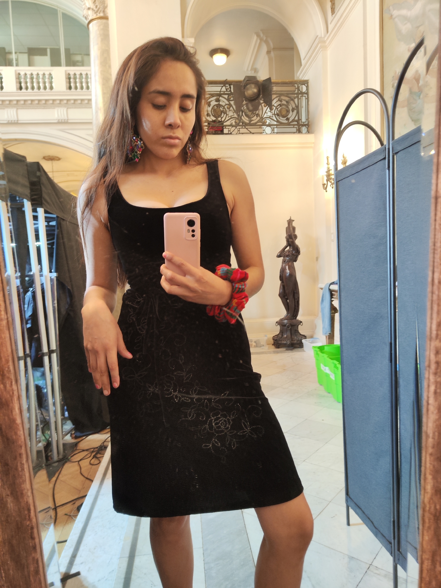 A young woman with long, dark brown hair is taking a mirror selfie in a well-lit indoor space, possibly a lobby or hallway, with polished marble floors. She is wearing a black sleeveless dress with a floral pattern and large, colorful beaded earrings. Her right hand is holding a pink smartphone, and a red and green scrunchie is around her wrist.
The background shows architectural details such as columns, ornate railings, and arched doorways, suggesting a grand building. To the left, part of a tall, dark draped object and some electrical equipment are visible in a mirror reflection. To the right, a folding screen with dark blue panels stands, and behind it, a green plastic bin and a dark statue of a person are reflected. The lighting appears to be a mix of ambient and artificial sources. The setting is reminiscent of the interior of the Museum of Fine Arts in Santiago, Chile, given the architectural style and the presence of art pieces like the statue, which aligns with the type of exhibits and the building's grandeur.