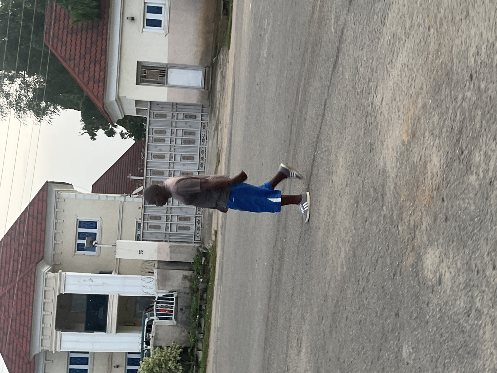 In Abuja, Nigeria, a man walks on a paved street. He is wearing a grey t-shirt and blue shorts. He has grey sneakers on his feet. Behind him is a beige building with a red roof and blue-trimmed windows. A large metal gate is visible in front of the building. There is also a smaller beige building with a red roof to the right of the larger building. Trees are visible in the background. The sky is overcast.