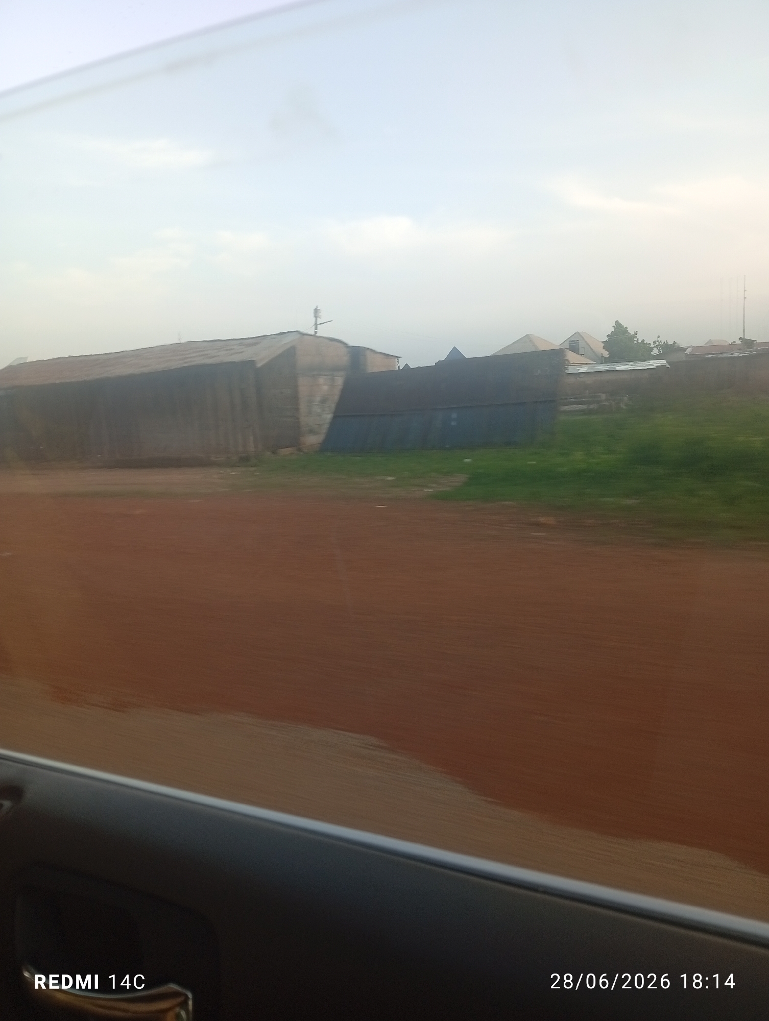 The image shows a street view of a residential area in Jos, Nigeria. The foreground features a dirt road, likely unpaved, with a slight blur indicating movement. Beyond the road, there are several buildings with corrugated metal roofs. Some of these structures appear to be simple dwellings or workshops. A patch of green grass is visible between the road and the buildings. The sky is overcast, with some lighter patches suggesting diffused sunlight. The overall impression is of a somewhat rural or developing urban landscape. The timestamp "28/06/2026 18:14" is embedded in the lower right corner, along with "REDMI 14C" in the lower left, indicating the device and date of capture.