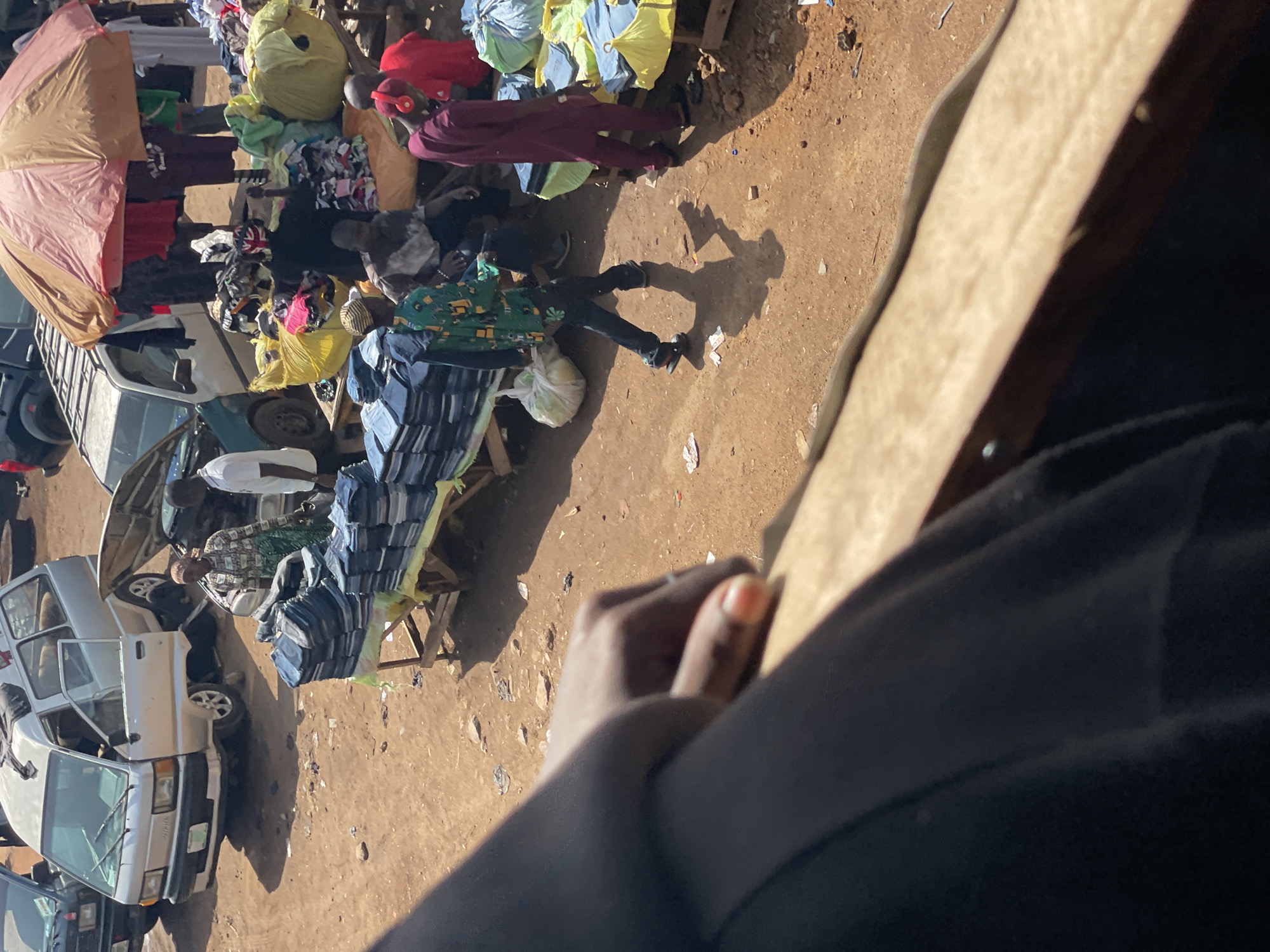 A bustling open-air market is depicted in this aerial shot, with various stalls and vehicles visible. In the foreground, a person's hand is resting on a wooden surface, partially obscuring the view of the market. The market appears to be selling clothing, with stacks of jeans and other garments displayed on tables and in bags. Several people are browsing and interacting at the stalls. A silver van with its doors open is parked in the background, and another vehicle with its hood up is also present. The ground is dirt, characteristic of an outdoor market. The scene suggests a lively commercial activity in a somewhat unpaved urban environment.
