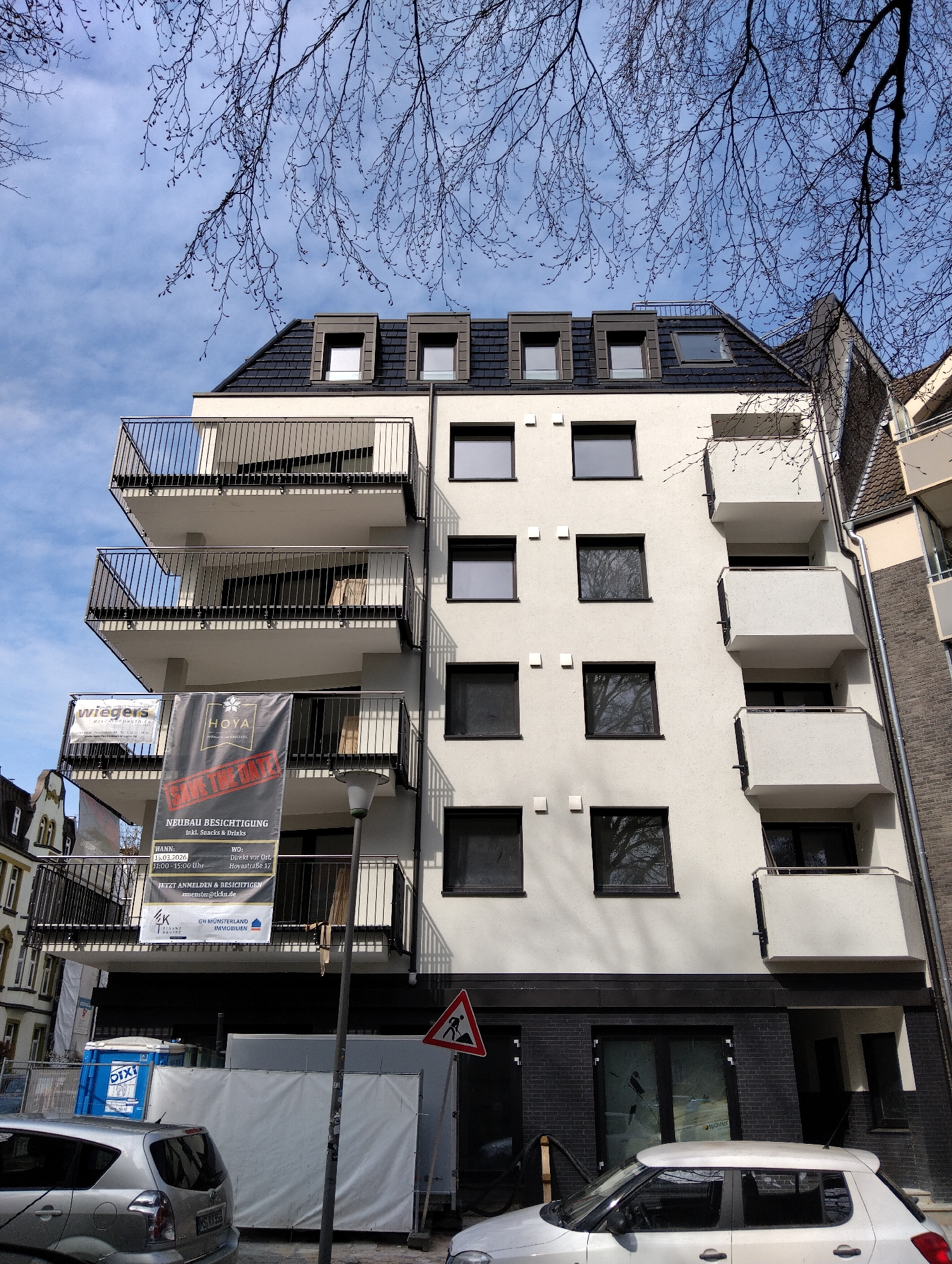 The image depicts a modern, multi-story residential building in Münster, Germany, as indicated by the German signage and the text "Hoyastraße 17." The building is white with black window frames and balconies. There are banners on one of the balconies advertising a "Neubau Besichtigung" (new building viewing) event scheduled for March 15, 2026. The banners feature the names "wiegers" and "HOYA." At the building's base, there's dark brick cladding, and signage with the text “isover” is visible behind what appear to be glass entryways. Construction or renovation activity is underway, evidenced by a portable toilet labeled "DIXI," construction barriers, and a warning sign indicating road work. Two parked silver/white cars are in front of the building. Branches from bare trees are visible in the upper portion of the image against a partly cloudy sky. The building is adjacent to another building, and a street lamp is also visible.