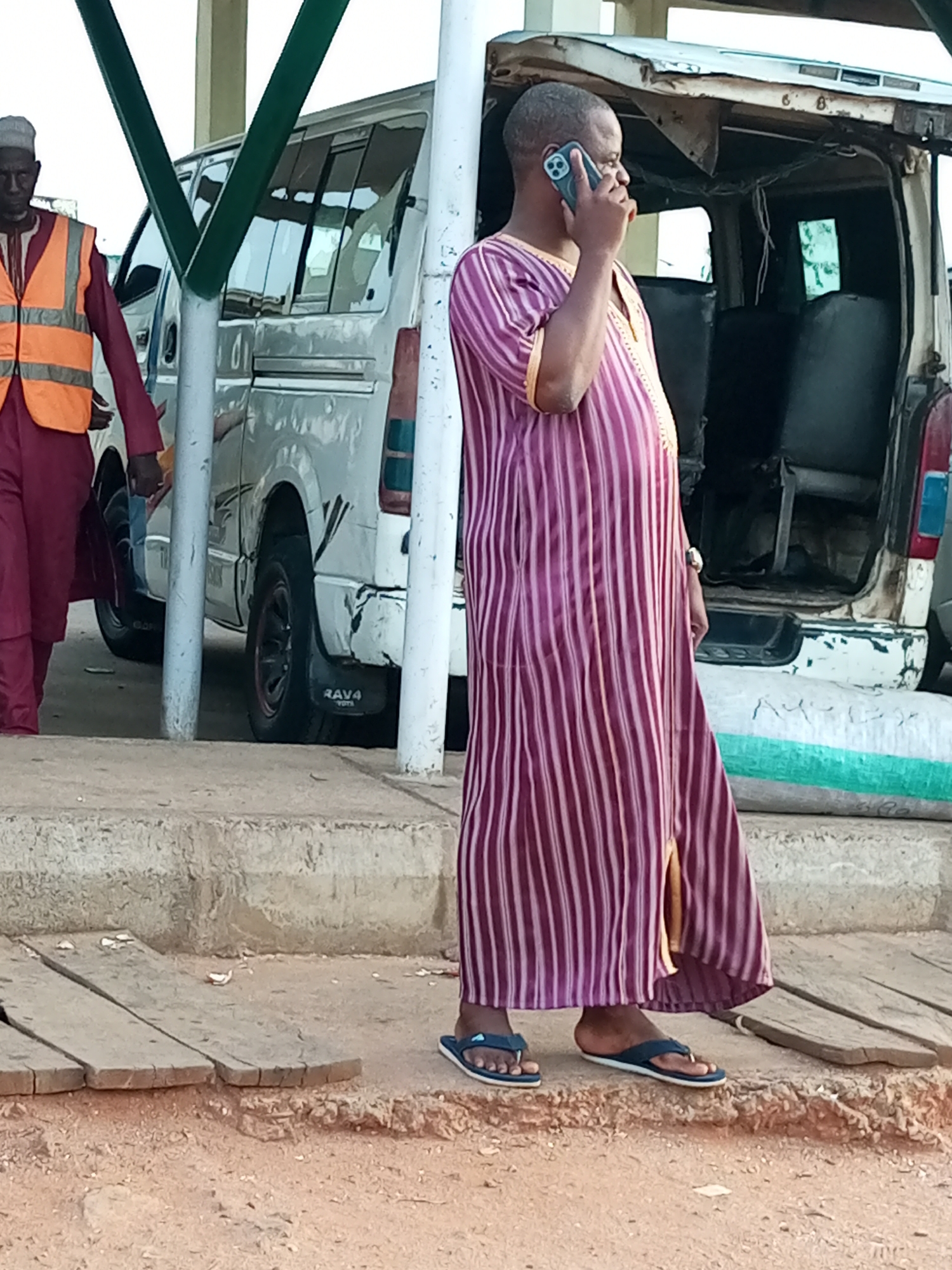 A man wearing a purple and white striped robe and dark sandals is standing and talking on a smartphone. He is positioned in the foreground, on a dirt and gravel surface with some wooden planks. Behind him, a white van with its rear doors open is parked on a paved area. To the left of the van, another man in a maroon outfit and an orange safety vest is walking. The scene appears to be outdoors, possibly a transportation hub or market area in Gombe, Nigeria, given the context of the van and the general surroundings.