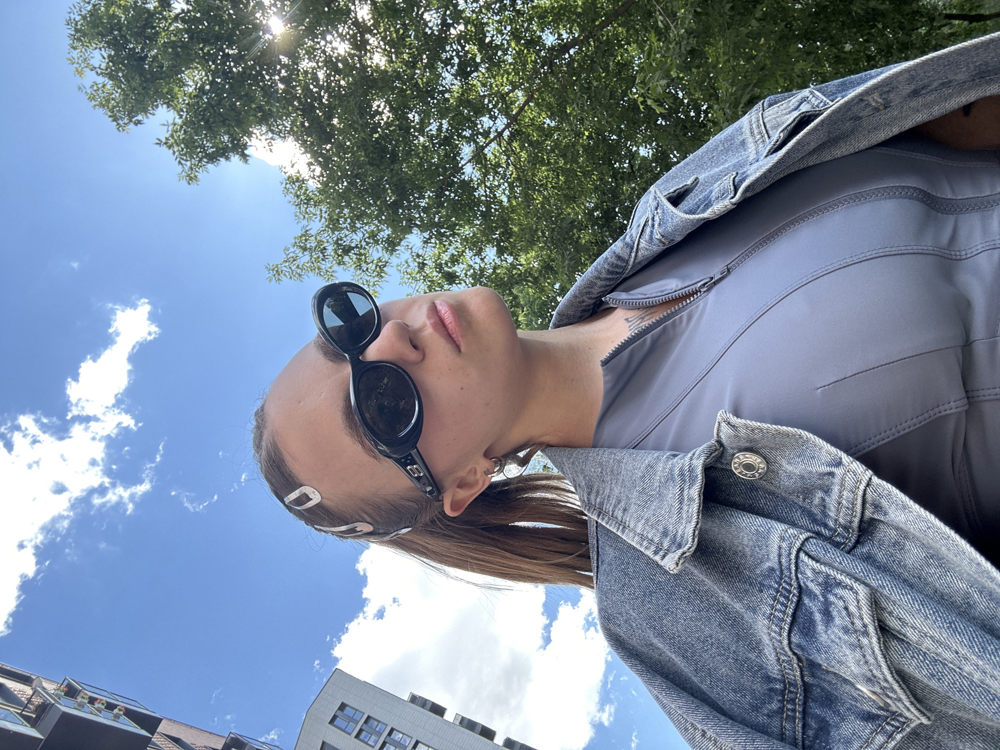 This media file is a close-up, low-angle shot of a young woman posing outdoors on a sunny day in Moscow, Russia. The woman is wearing a denim jacket over a light grey top, and large black sunglasses. Her head is tilted back, looking up at the bright blue sky with some scattered white clouds. Lush green trees with dappled sunlight shining through them are visible in the upper portion of the frame. In the background, the tops of modern buildings can be seen. The lighting suggests it is daytime, likely late morning or early afternoon. The woman's expression is neutral. There are no visible text elements in the image. The overall impression is one of casual outdoor activity on a pleasant day.