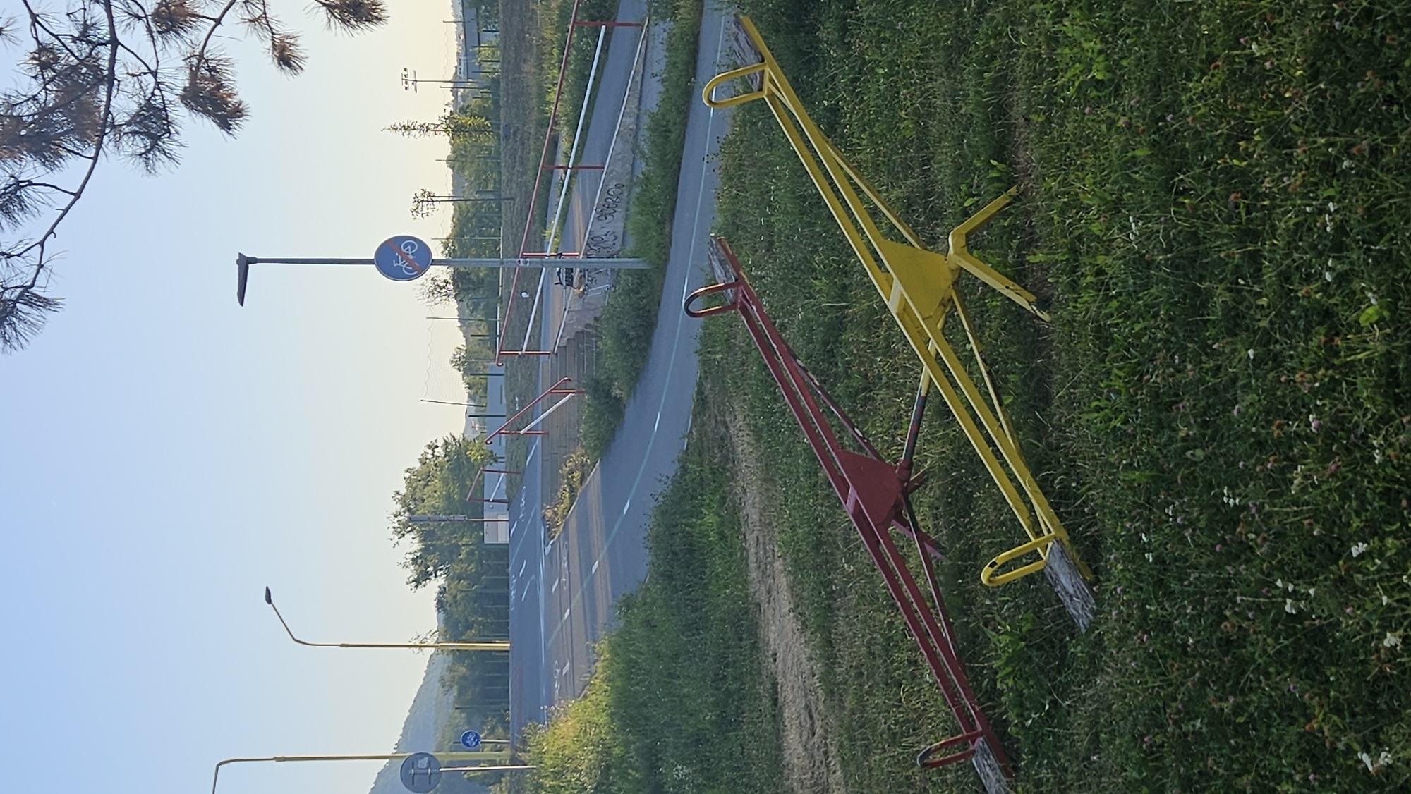 The image depicts a park area in Prešov, Slovakia, featuring two seesaws in the foreground. One seesaw is red, and the other is yellow, both situated on a grassy terrain dotted with small white and pink flowers. In the background, a paved road with painted lane markings curves upwards. Red and white railings line the side of the road. Further in the distance, there are streetlights and various trees. A sign with a bicycle symbol crossed out is visible on a pole. The scene is illuminated by natural daylight, suggesting it is daytime.