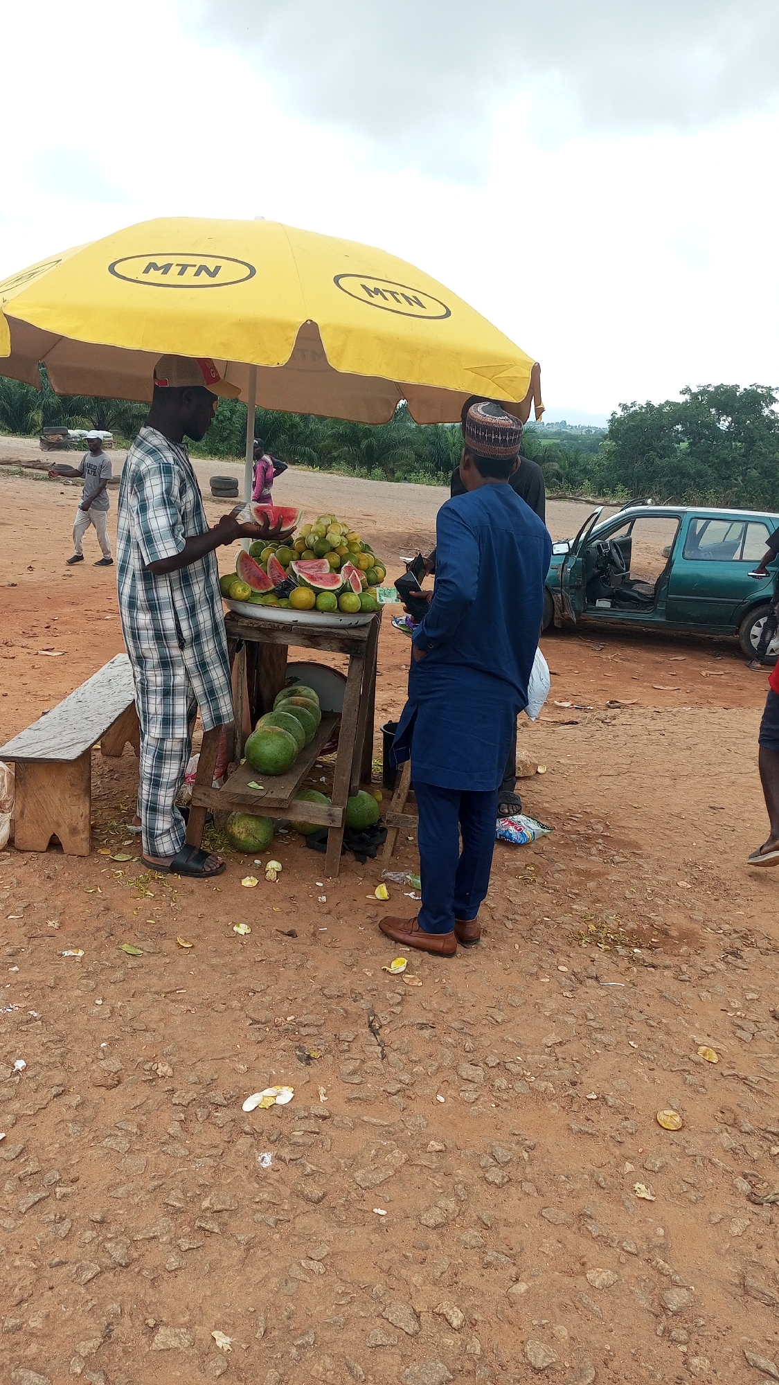 In Dauda, Nigeria, two men stand by a roadside fruit stand under a large yellow umbrella bearing the "MTN" logo. One man, wearing a checkered shirt and pants, is in the process of selecting a slice of watermelon from a bowl of assorted fruits. The other man, dressed in a blue tunic and pants, faces away from the viewer, observing the fruit selection. A wooden stand displays several watermelons on a lower shelf and a variety of fruits, including oranges and watermelons, on an upper shelf. In the background, a green vehicle with an open door is parked nearby. The ground is covered in dirt and scattered fruit peels. The scene appears to be a typical roadside vendor in a rural or semi-urban setting.