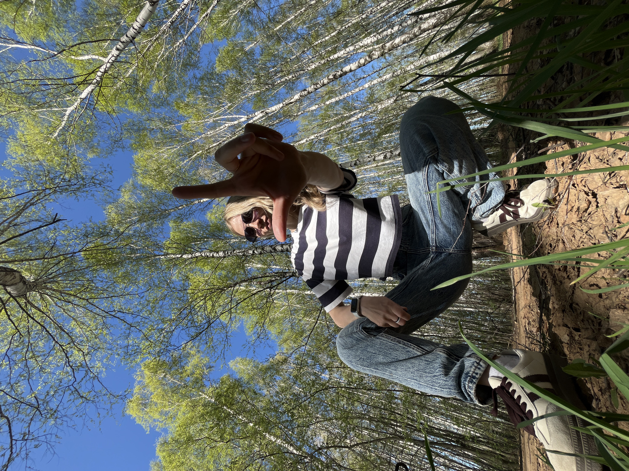 A woman wearing jeans, a striped shirt, sunglasses, and sneakers is crouched in a forest. She is holding her hand up to the camera. The trees are birch trees, and the sky is blue. Tall grass is visible on the right side of the image. The location is Chaikovsky, Russia.