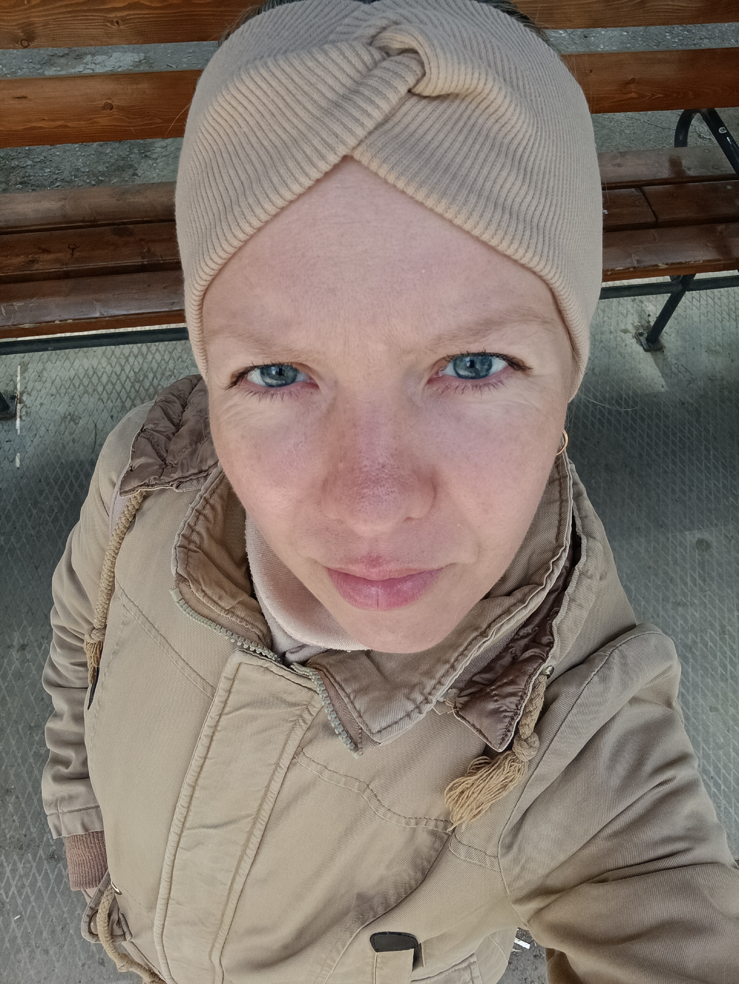 A woman with light skin and blue eyes is looking up at the camera. She is wearing a beige, twisted turban-style hat and a beige jacket with brown quilted trim around the collar. The jacket is zipped up. She has a small gold hoop earring in her left ear. The background consists of a wooden bench above and a gray, patterned surface below. The photo was likely taken outdoors during the day. The city of Chaikovsky, Russia, is not discernible in the image.