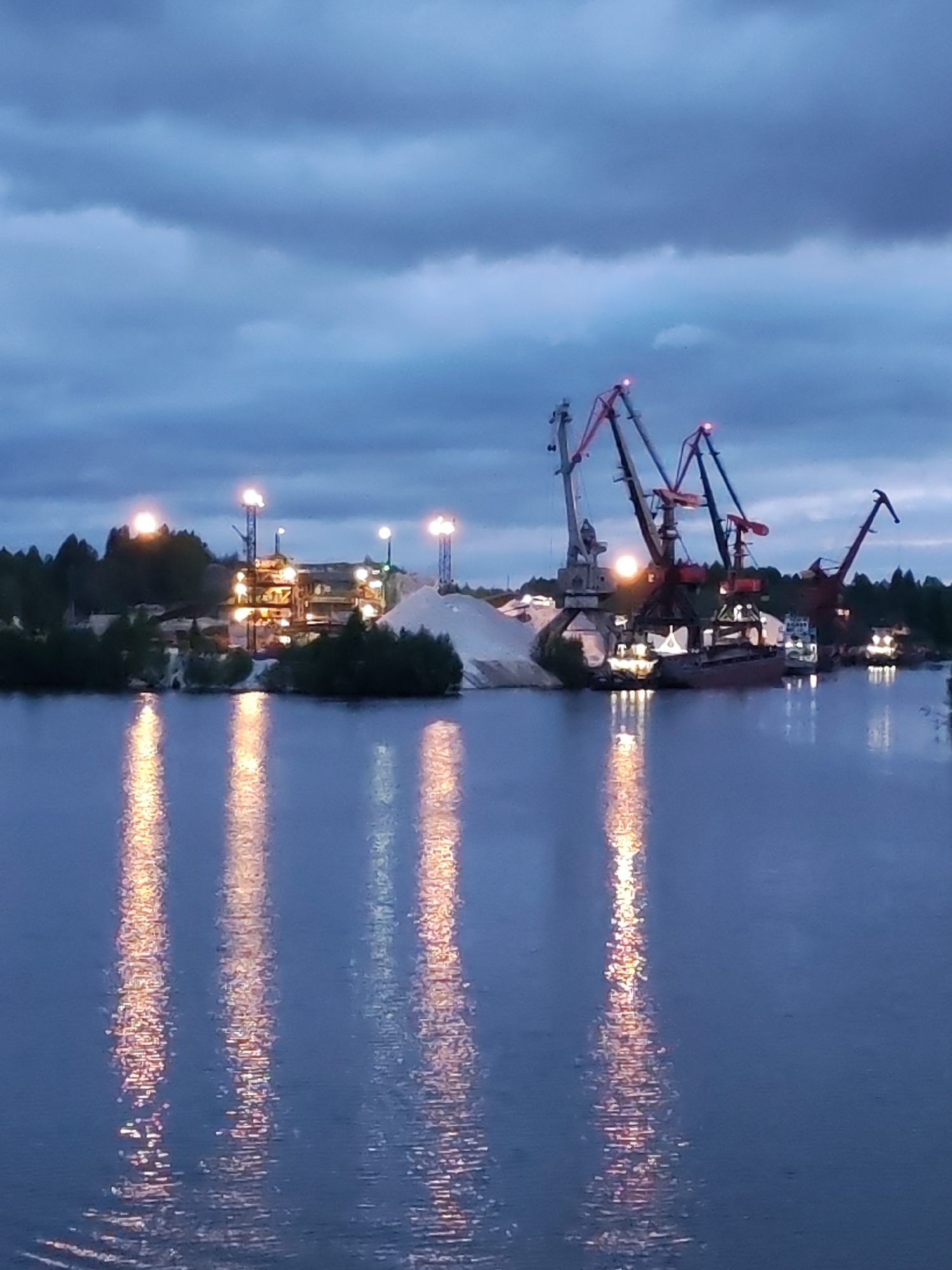 The image shows an industrial scene at dusk in Старое Петровское, Russia. The foreground is dominated by a body of water reflecting the lights from the structures on the shore. In the midground, there's a complex of buildings and a large pile of materials, possibly sand or gravel, illuminated by several lights. Four cranes, including two red, one grey, and one dark red, are positioned along the waterfront. A barge is docked next to the cranes. The background features a line of trees against a cloudy, blue-grey sky.