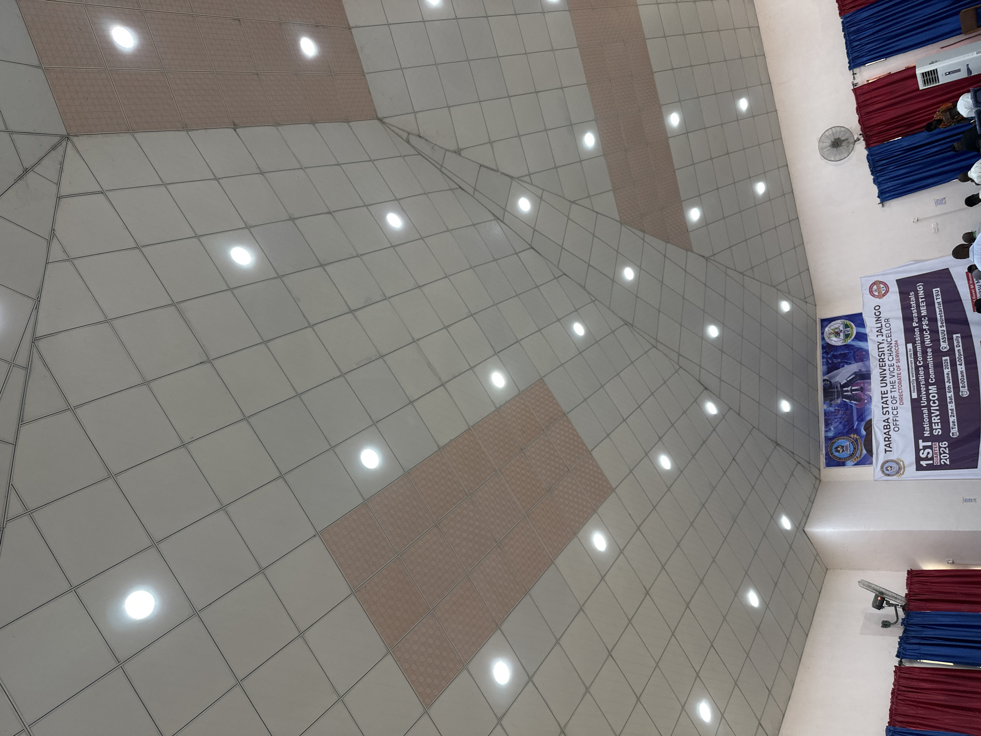 The image shows the interior of a large room with a domed ceiling. The ceiling is made of square tiles, with some panels featuring a diamond pattern. Numerous circular lights are embedded in the ceiling. In the foreground, a banner is partially visible, indicating a "1ST National Universities Commission Parastatals SERVCOM Meeting" in 2026, held in Jalingo, Nigeria. Red and blue curtains hang on the walls, and an industrial fan is mounted on the wall. Several people are seated or standing in the background. The overall impression is of an indoor event or meeting space.