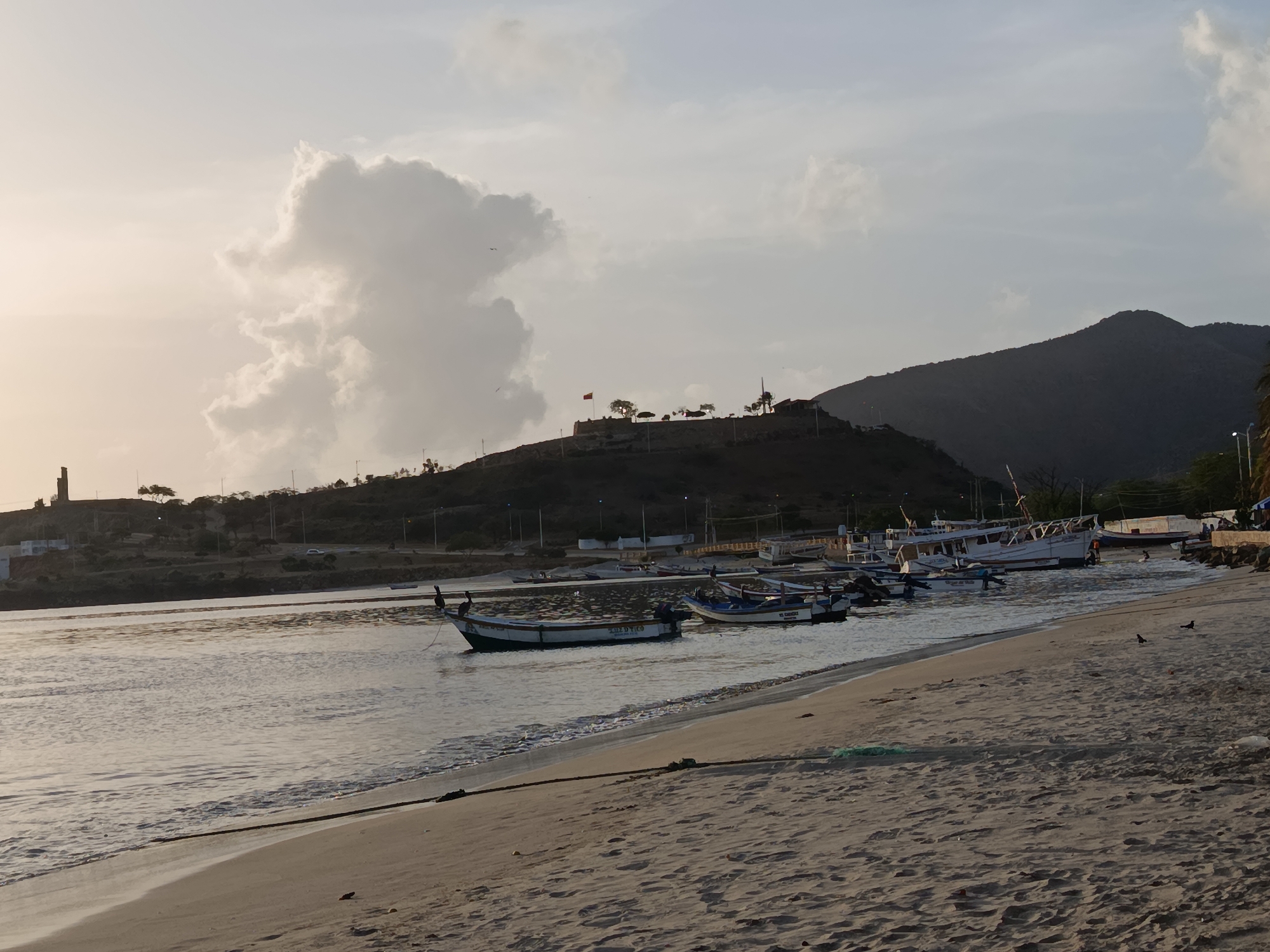 The image depicts a sunset scene at the bay of Juan Griego, Venezuela. The foreground shows a sandy beach with a rope stretched across it. The calm water of the bay is filled with several small fishing boats, some with motors. In the middle ground, a hill rises with a structure on top, possibly a fort or historical site, marked by a red flag. The background is dominated by a large, dark mountain. The sky is partly cloudy, with a large, luminous cloud formation on the left side. The lighting suggests it is late afternoon or early evening.