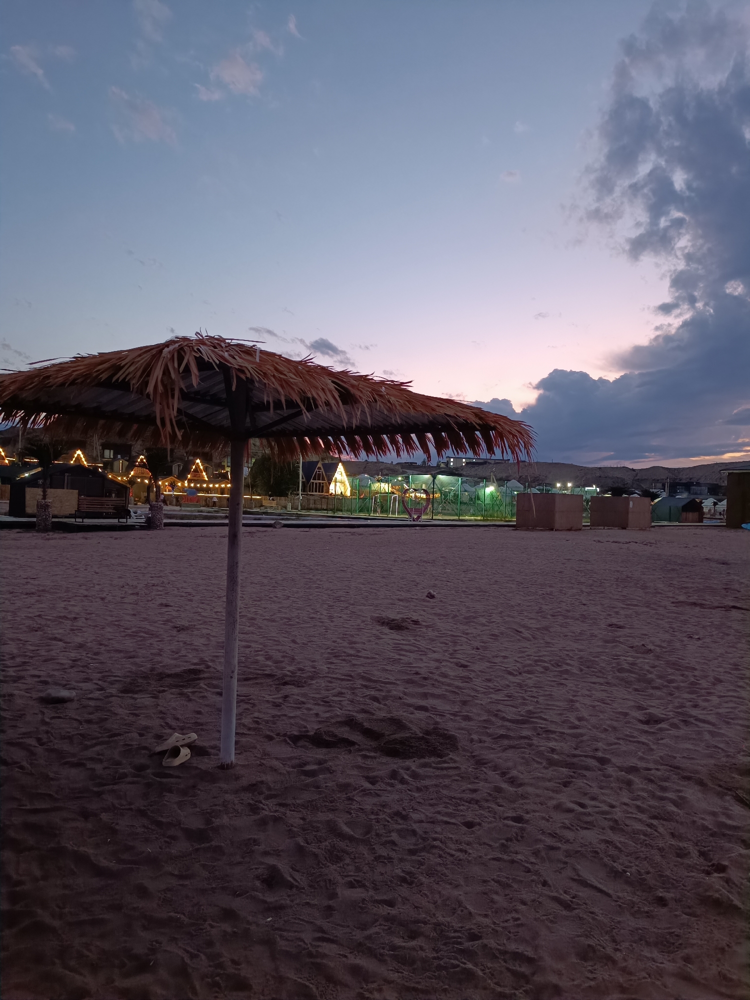 This is a vertical, low-angle, outdoor photograph taken at dusk on a beach. The foreground is dominated by dark, sandy beach with footprints. A light-colored pole supports a large, thatched umbrella in the center of the frame. To the left of the umbrella, a pair of light-colored flip-flops rests on the sand.
In the mid-ground, a row of small, illuminated structures resembling huts or cabins are visible. They are decorated with strings of warm, yellow lights, creating a festive atmosphere. A bench is also visible to the left of the structures. A fence with green netting runs along the front of these structures. Two large, cardboard boxes are placed on the sand to the right of the structures.
The background features a hazy hillside with some lights scattered across it. The sky is a gradient of twilight colors, transitioning from pale blue at the top to soft pinks and purples near the horizon. Dark, heavy clouds are present in the upper right portion of the sky.
The image appears to depict a beachside resort or recreational area, possibly at Issyk-Kul Lake in the city of Tong, Kyrgyzstan, given the presence of the lake and the general aesthetic. There are no people visible in the frame. The overall mood is serene and peaceful, with a hint of anticipation for the evening.