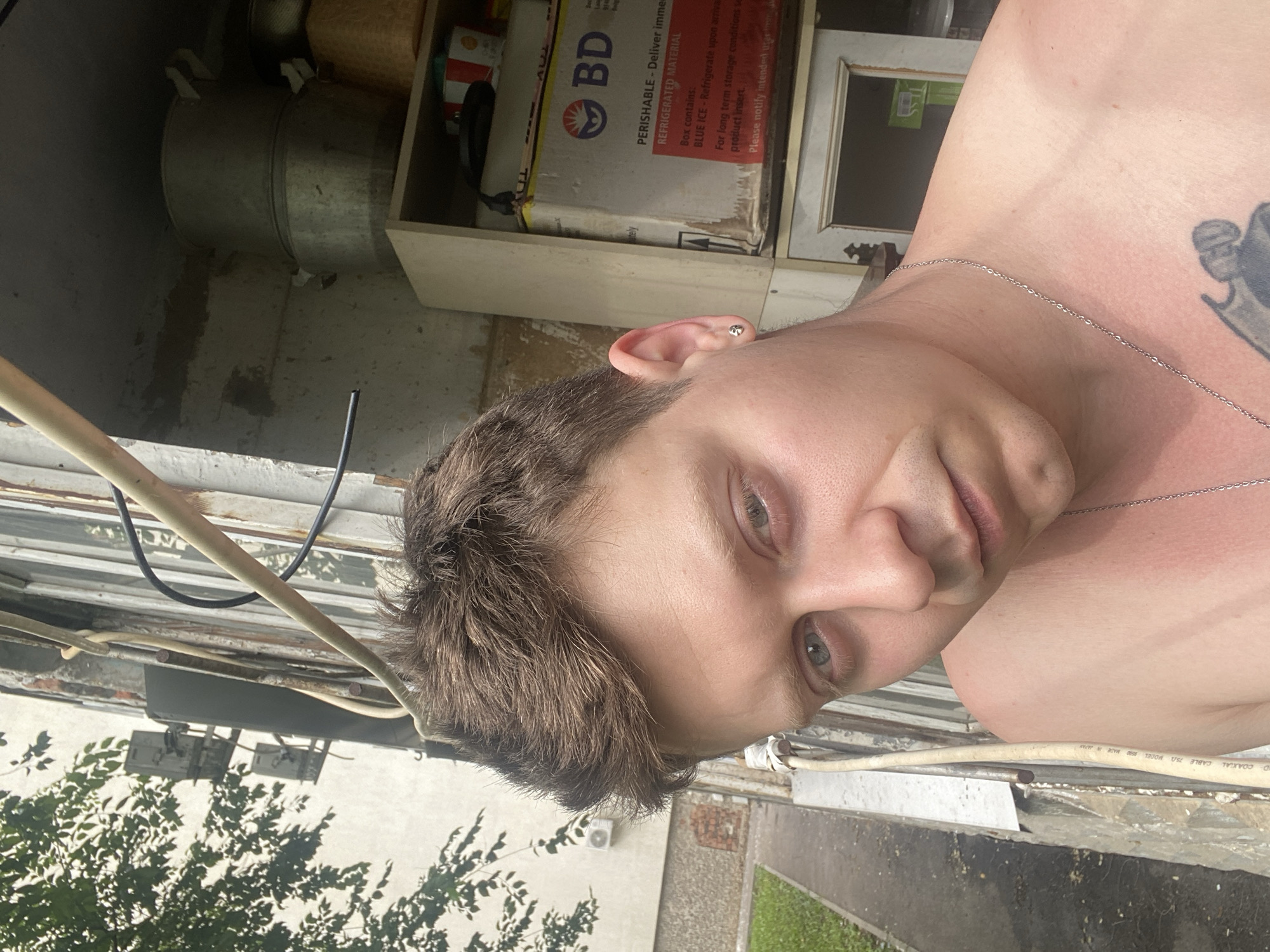 A young man with brown hair and blue eyes is pictured from the chest up, looking directly at the camera. He is wearing a silver chain necklace and has a small stud earring in his left ear. A tattoo is visible on his upper chest. The background suggests an urban setting, likely a residential building with exposed pipes and electrical conduits running along the exterior wall. A tree with green leaves is visible on the left, and a metal railing is present in the foreground. A shelf unit containing various items, including boxes and containers, is located behind him. The lighting indicates it is daytime, with the sun casting shadows. The image appears to be a selfie taken outdoors.